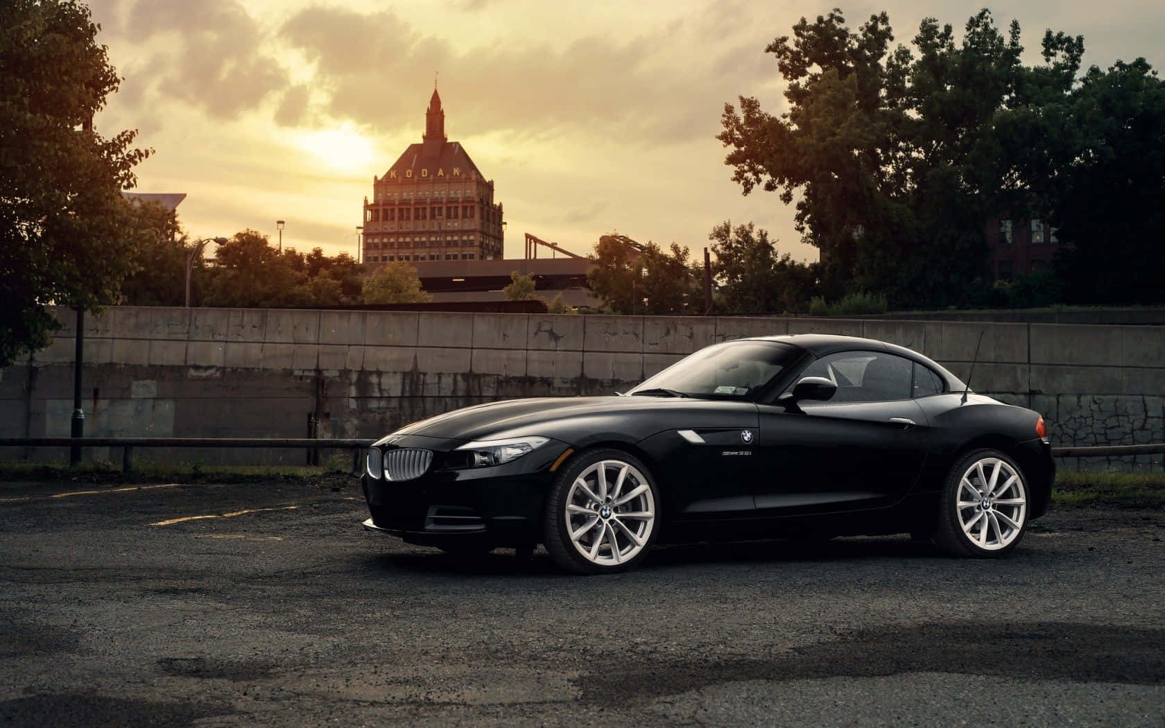 Bmw Z4 桌布 壁紙 — 一輛時尚的黑色BMW Z4跑車停泊在日落時分的城市空地，背景有歷史建築和樹木。