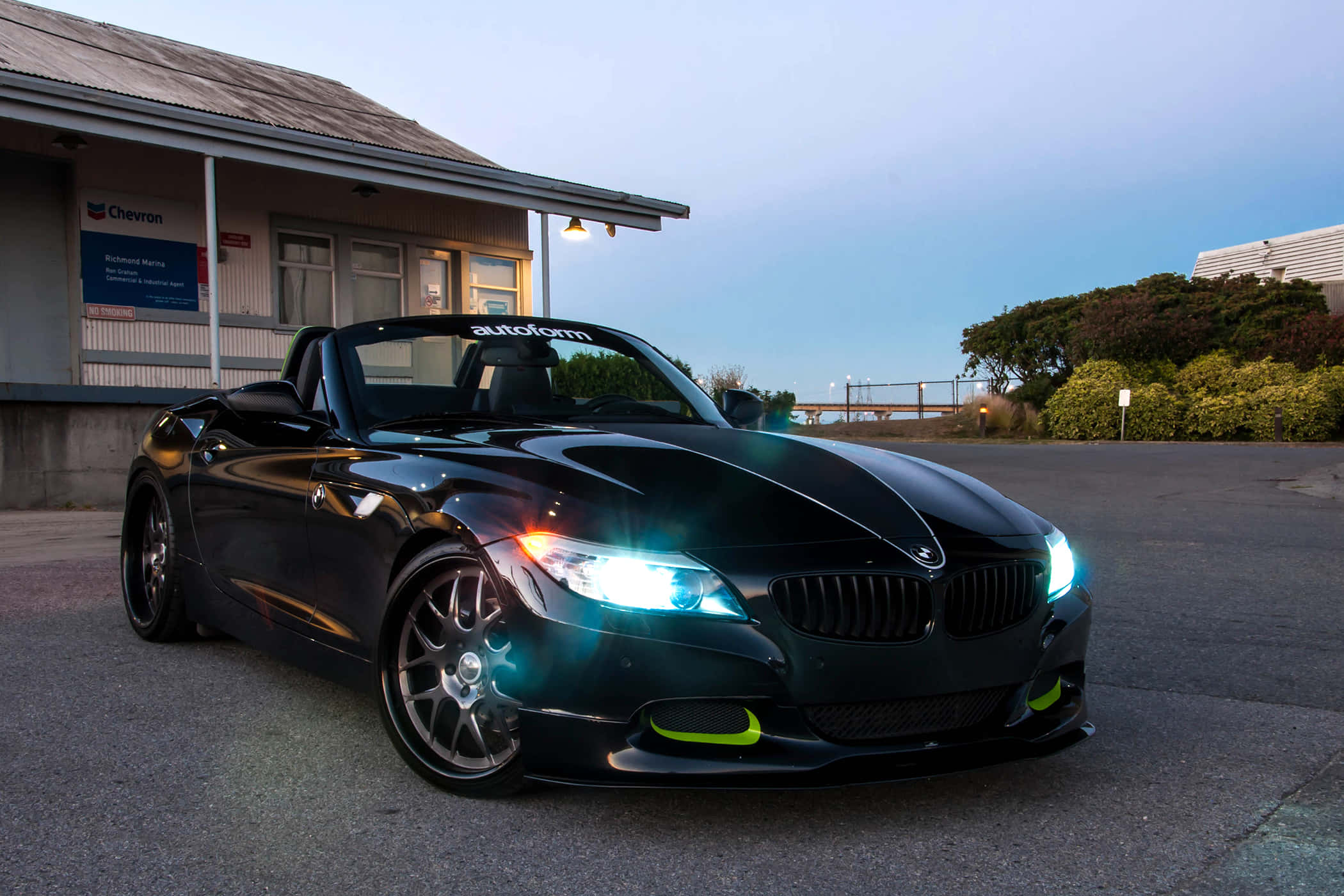 Bmw Z4 桌布 壁紙 — 一輛時尚的黑色BMW Z4開篷跑車，車頭燈閃爍，停泊在雪佛龍加油站前，正值黃昏。