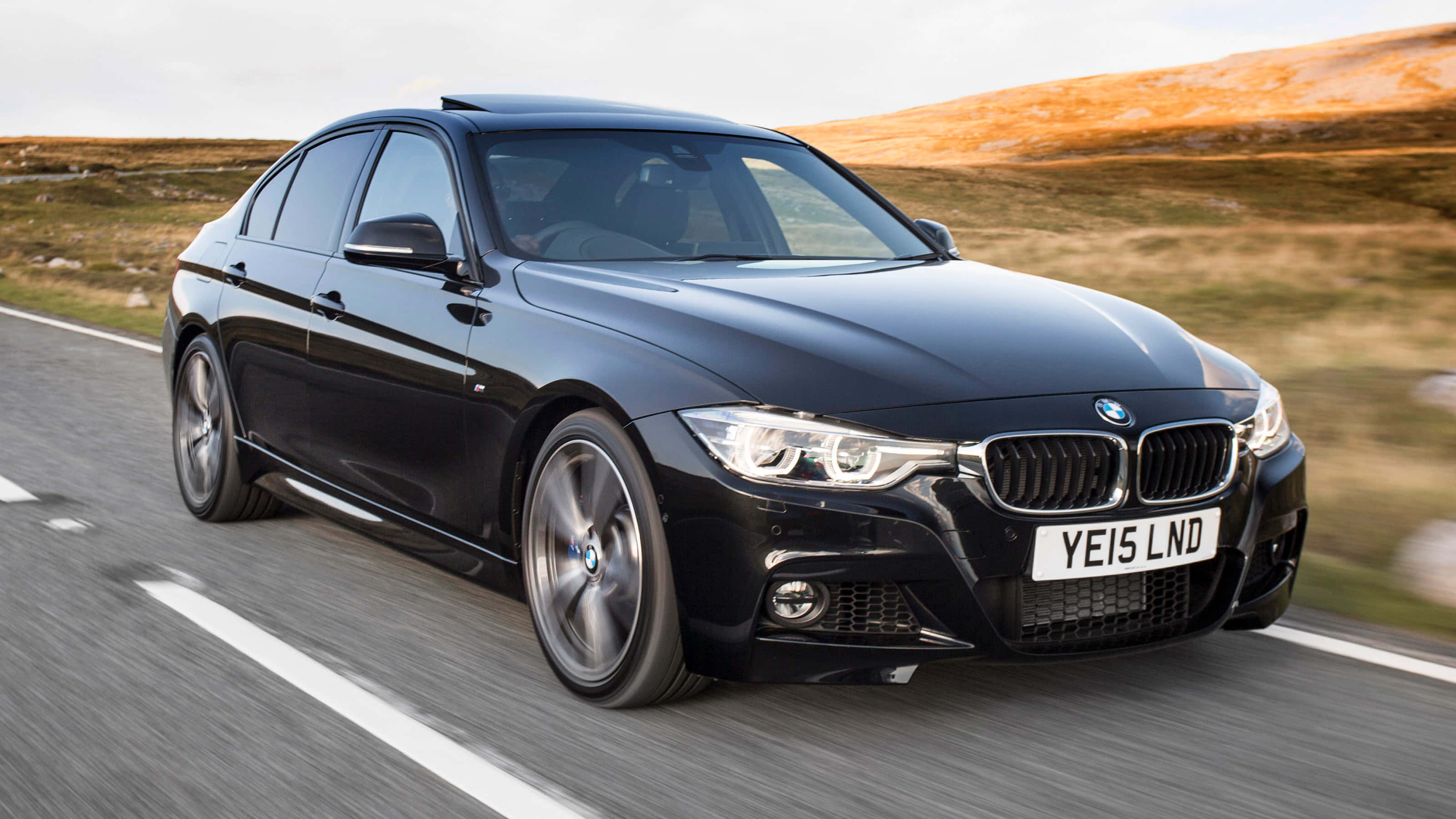 Bmw 3 Series 桌布 壁紙 — 一輛時尚的黑色BMW房車在晴朗天空下，沿著蜿蜒的道路穿過起伏的金色山丘飛馳。