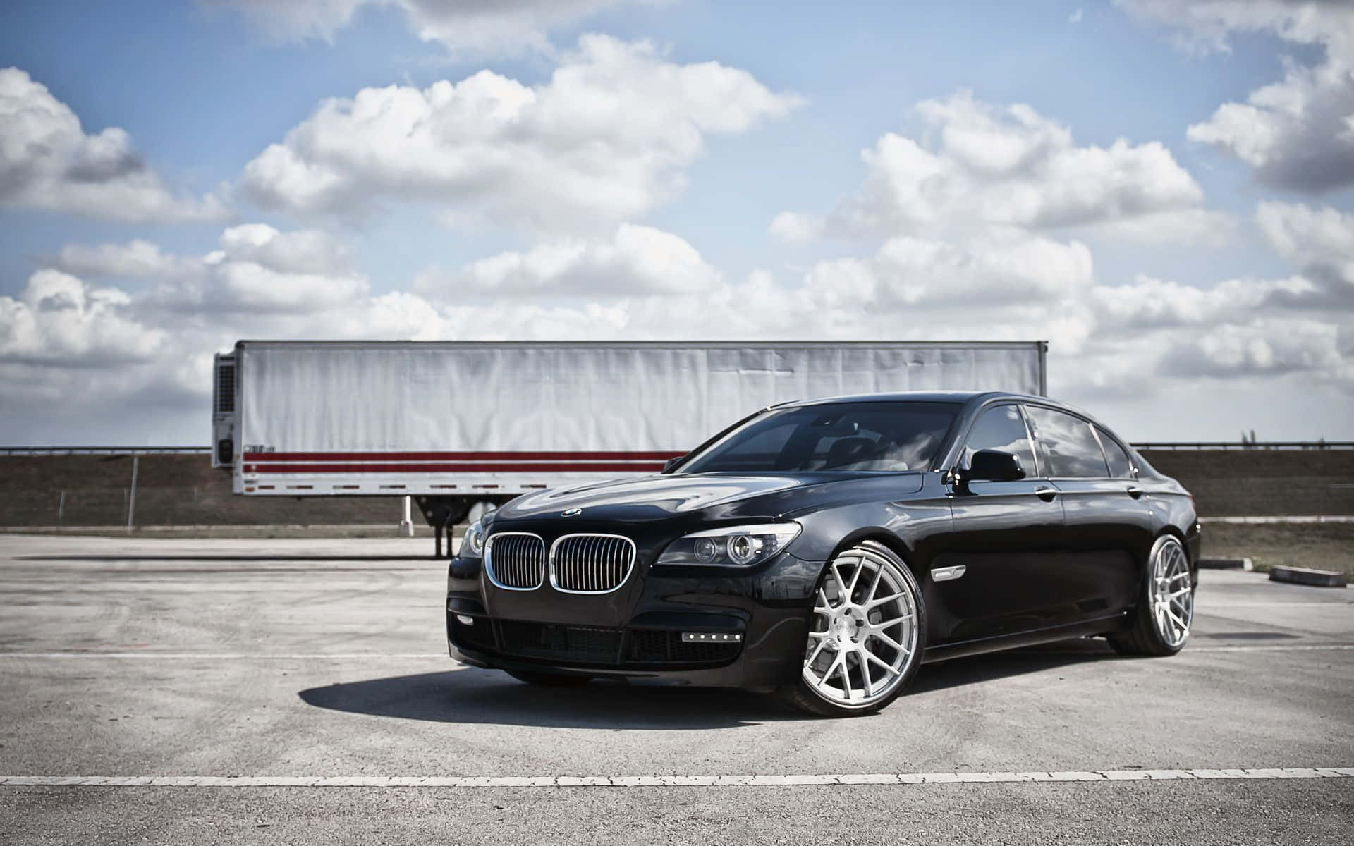 Bmw 7 Series 桌布 壁紙 — 一輛時尚的黑色BMW轎車，配備定制銀色輪圈，停在瀝青路面上，背景是明亮的多雲天空和一輛白色半掛卡車。