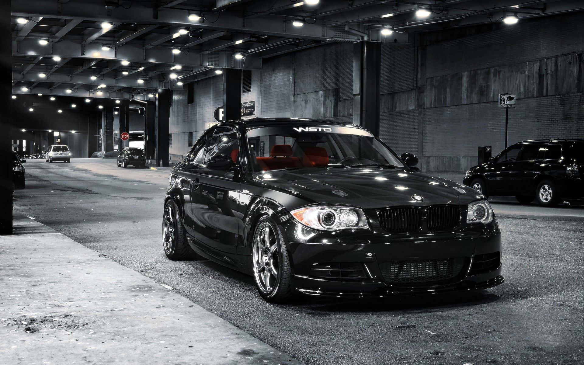 Bmw 1 Series 桌布 壁紙 — 一輛時尚的黑色BMW雙門轎跑車，紅色內飾，停在光線昏暗的地下停車場，有混凝土牆和頭頂燈光。