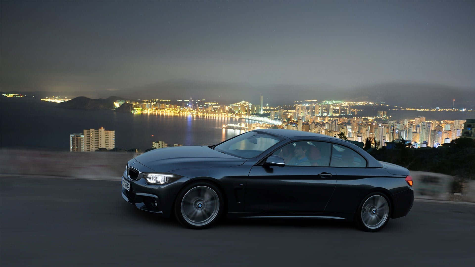 Bmw 4 Series 桌布 壁紙 — 一輛時尚的黑色BMW雙門轎跑車沿著海岸公路夜間行駛，背景是閃爍的城市天際線和港口燈光。
