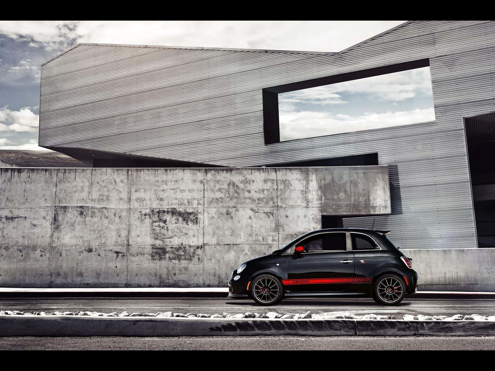 Abarth 桌布 壁紙 — 一輛時尚的黑色Fiat 500，帶有紅色點綴，停在一棟現代混凝土建築旁邊，天空多雲。