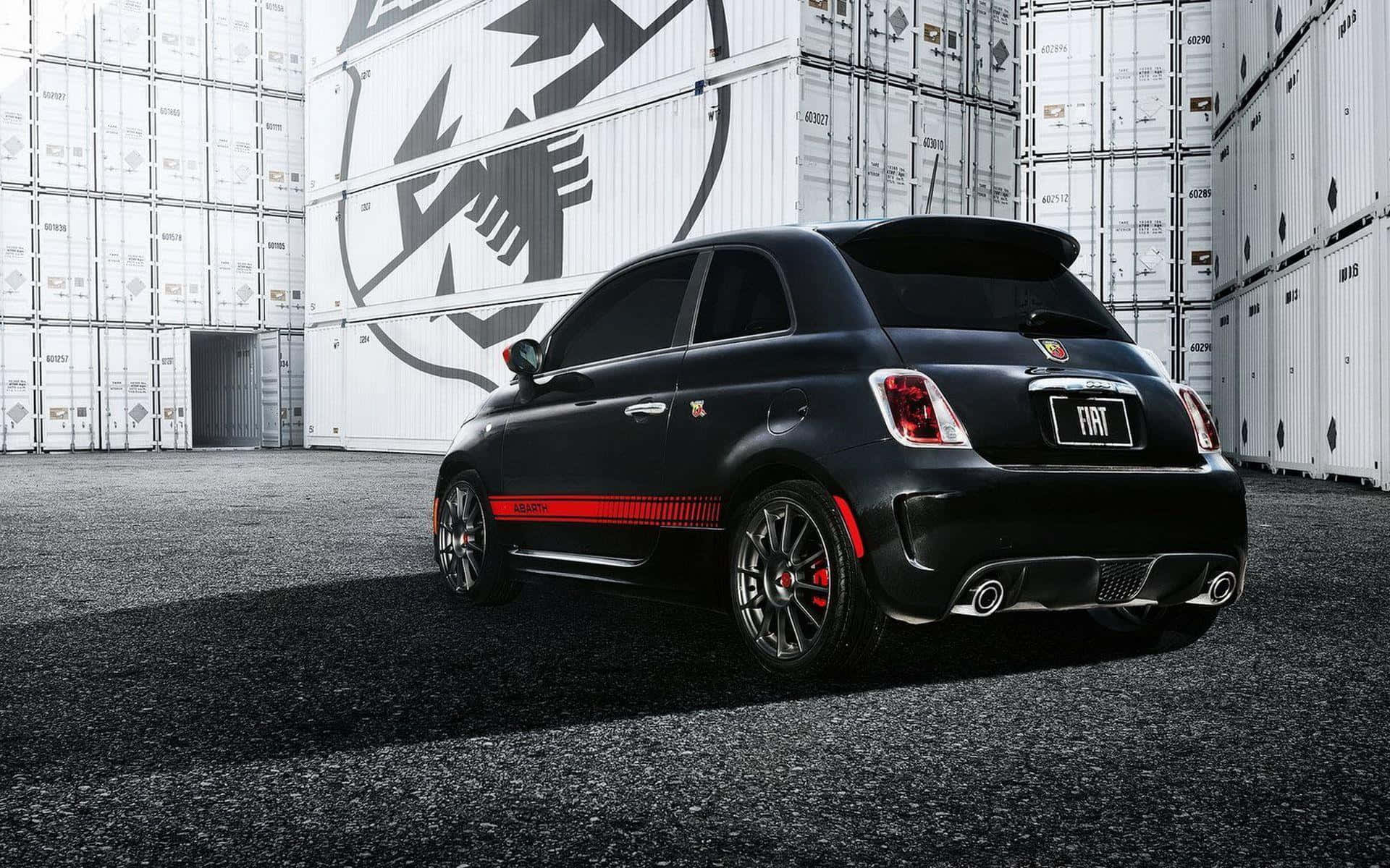 Abarth 桌布 壁紙 — 一輛時尚的黑色Fiat 500帶有紅色點綴，停在堆疊的白色貨櫃之間，在明亮的日光下。