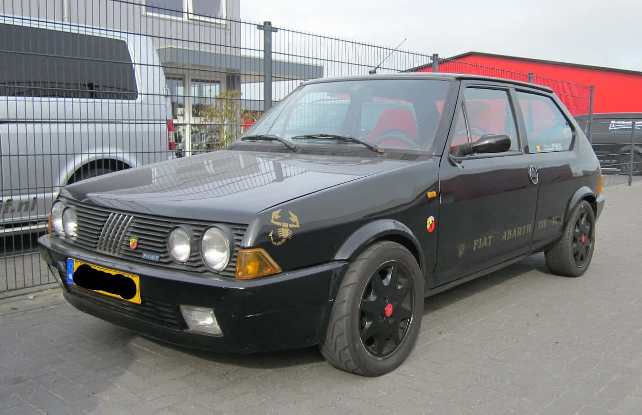 Fiat Ritmo 桌布 壁紙 — 一輛時尚的黑色Fiat Abarth 130 TC跑車停在行人路上，配備紅色內飾和金色蠍子標誌。