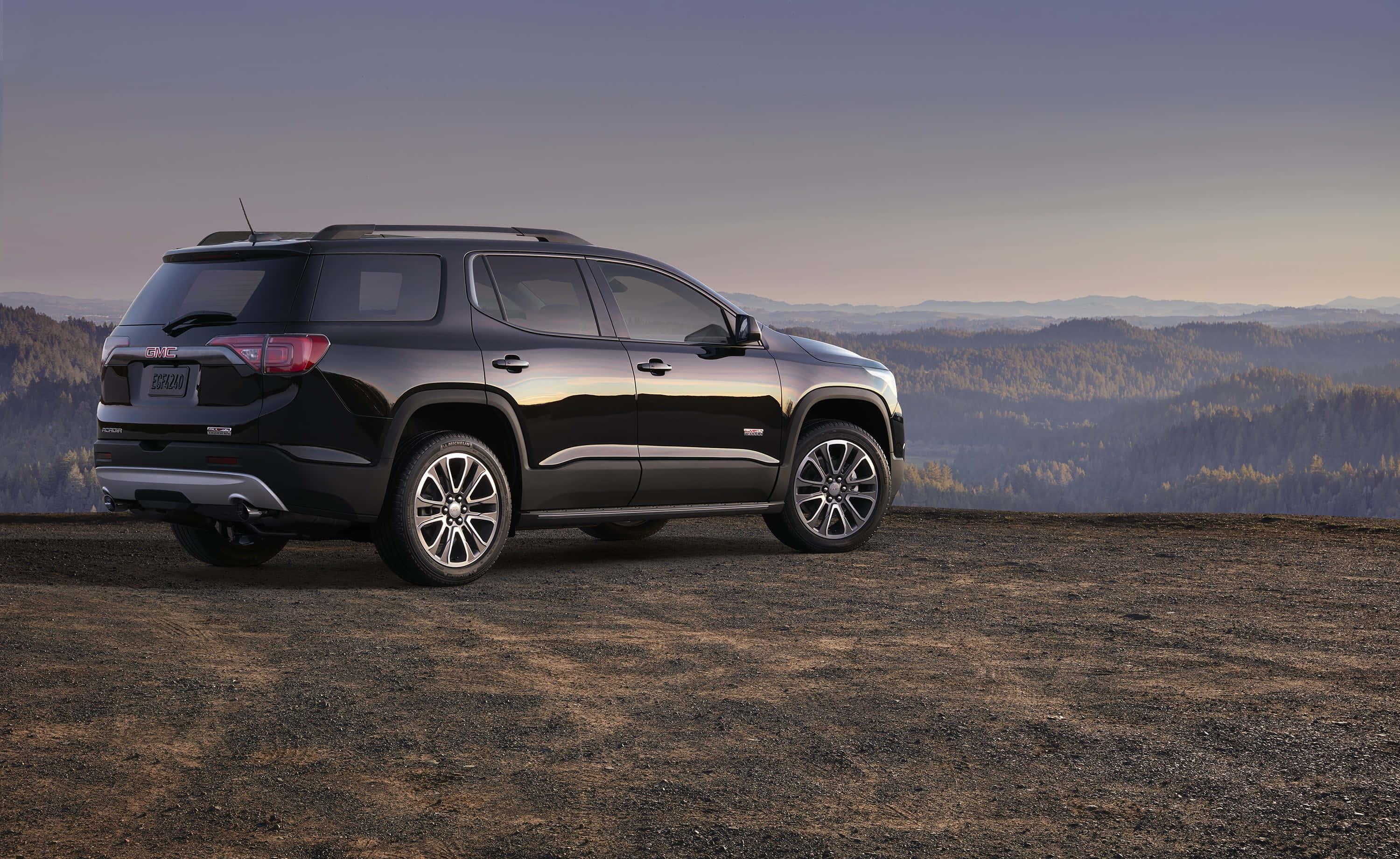 Gmc Acadia 桌布 壁紙 — 一輛時尚的黑色GMC Acadia SUV停泊在碎石觀景點上，連綿起伏的森林山丘在柔和暮色天空下逐漸消失在霧氣瀰漫的遠方。