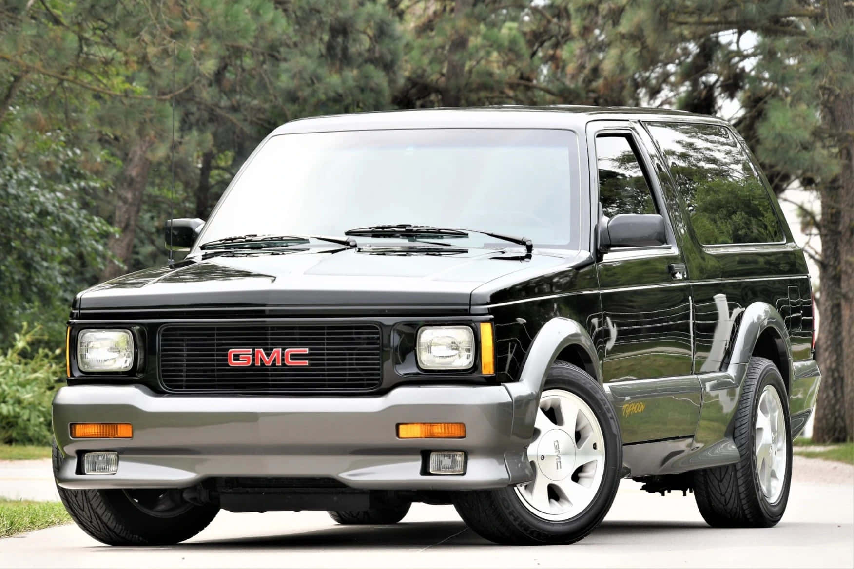Gmc Typhoon 桌布 壁紙 — 一輛時尚的黑色GMC Safari SUV停泊在鋪砌的道路上，背景有樹木。