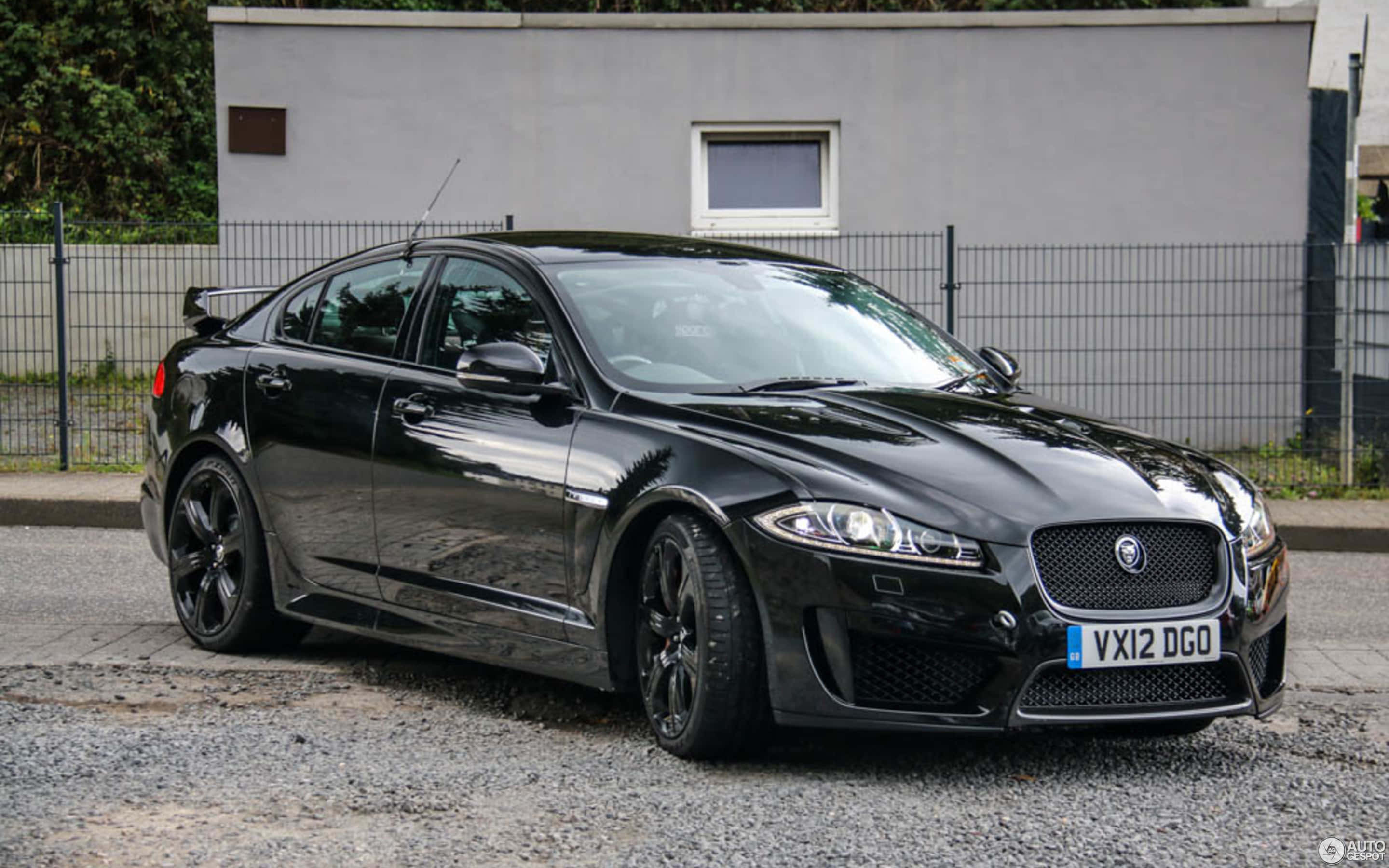 Jaguar Xfr 桌布 壁紙 — 一輛時尚的黑色Jaguar XFR運動轎車停在碎石上，後面有灰色牆壁和圍欄。