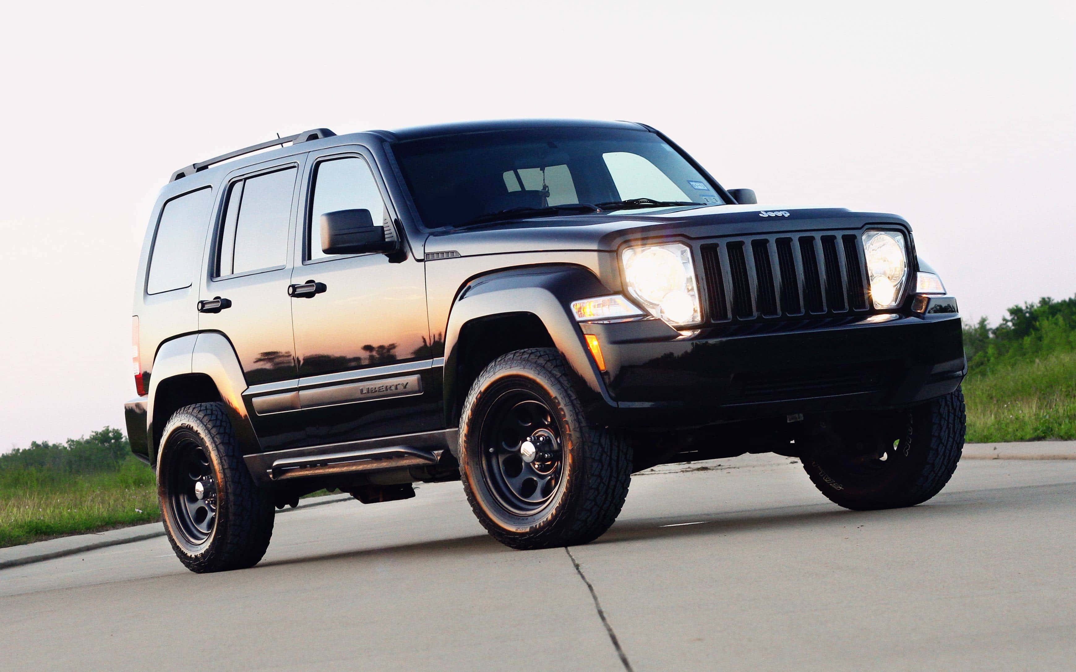 Jeep Liberty 桌布 壁紙 — 一輛時尚的黑色Jeep Liberty SUV，車頭燈閃爍，停在日落時分的瀝青路上，背景是草地田野。