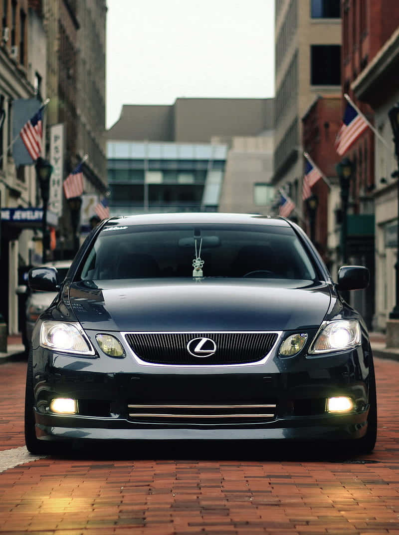 Lexus Gs 桌布 壁紙 — 一輛時尚的黑色Lexus轎車在鋪滿磚塊的城市街道上向前行駛，街道兩旁是建築物和美國國旗。