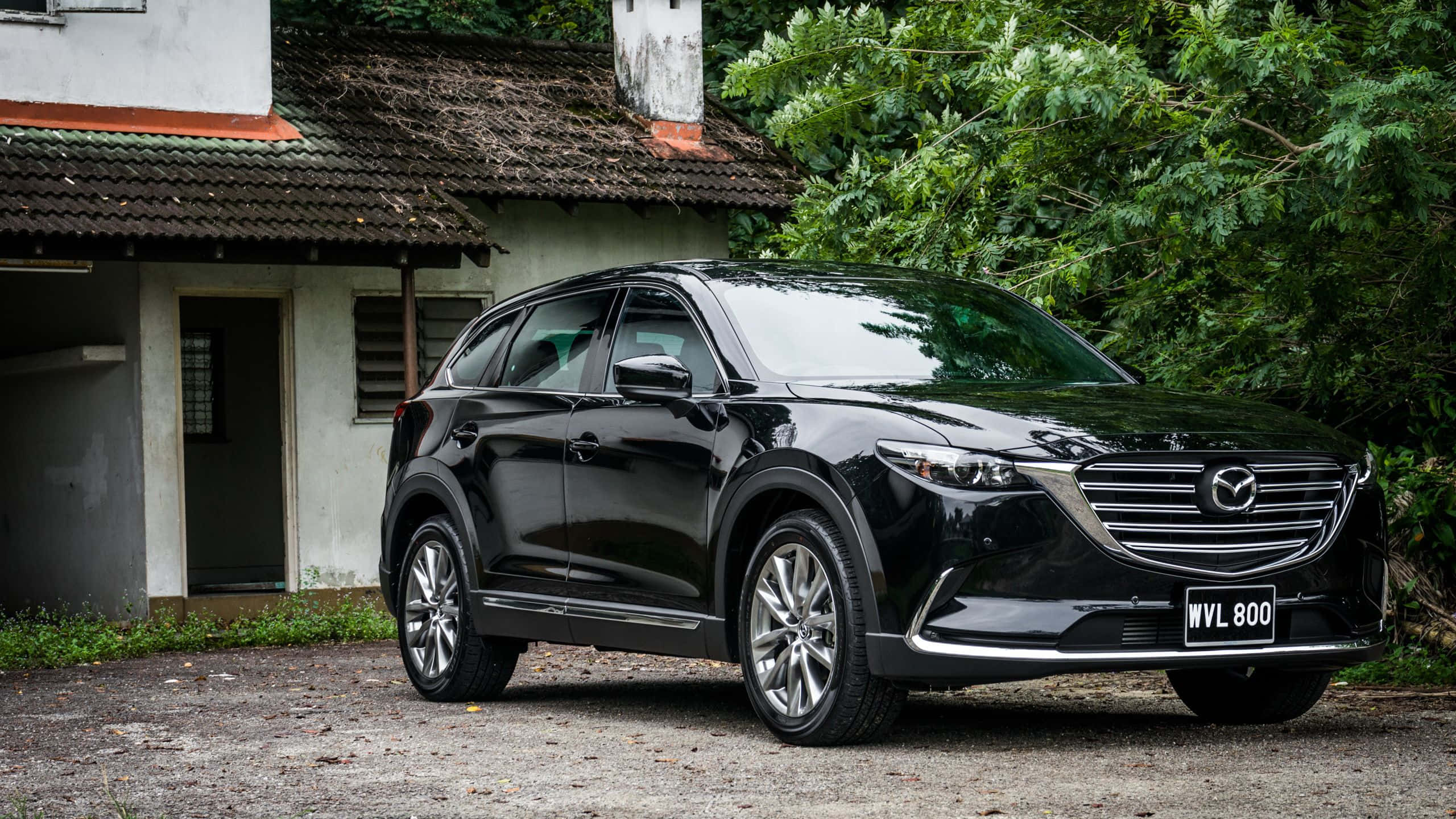 Mazda Cx-9 桌布 壁紙 — 一輛時尚的黑色Mazda CX-9 SUV停泊在碎石路上，前面是一間有茂密綠色植物的鄉村風格房屋。