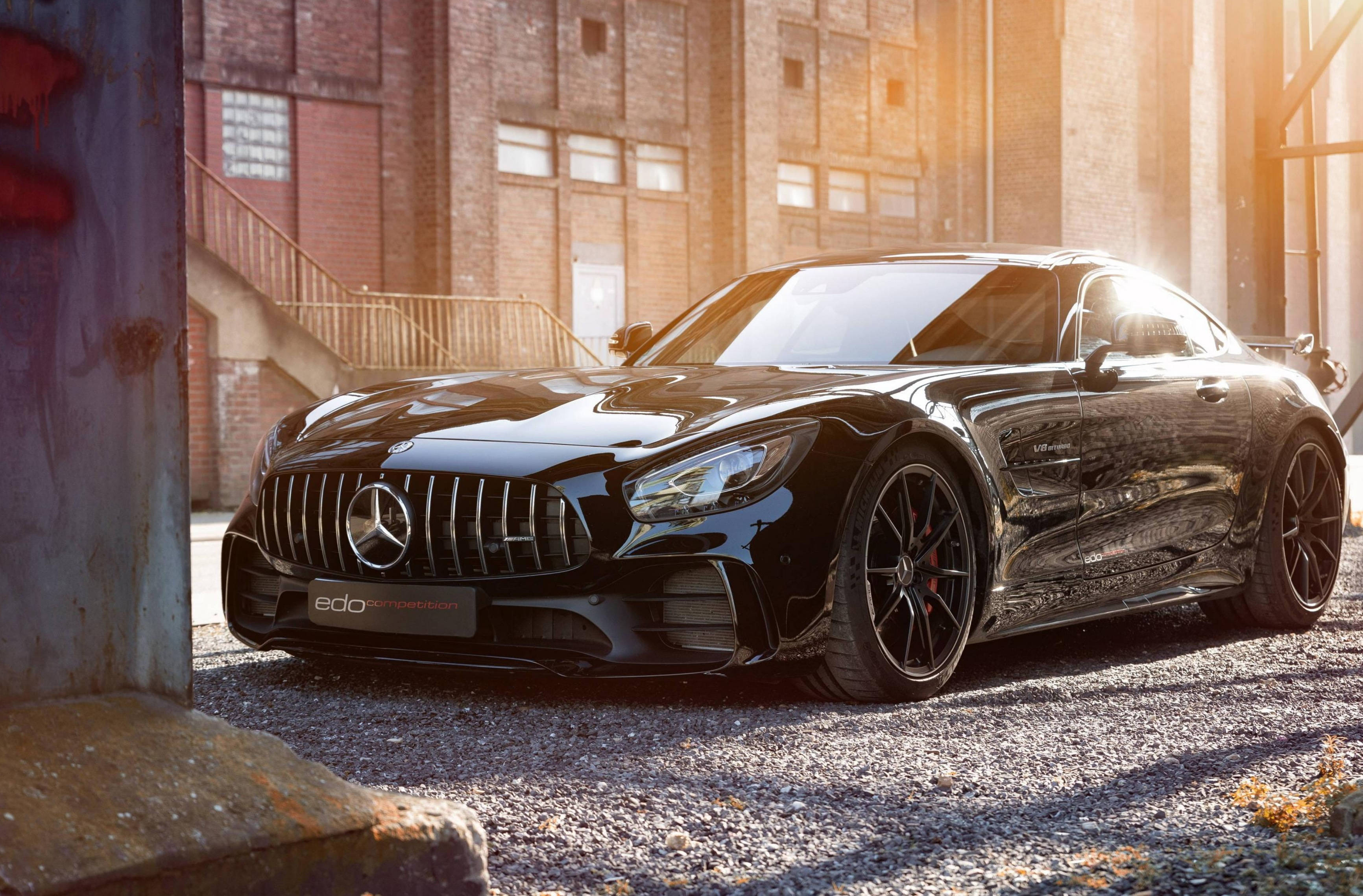 Mercedes Benz 桌布 壁紙 — 一輛時尚的黑色Mercedes-AMG GT停在碎石路上，前方是陽光普照的磚造倉庫，溫暖的黃金時刻光線。