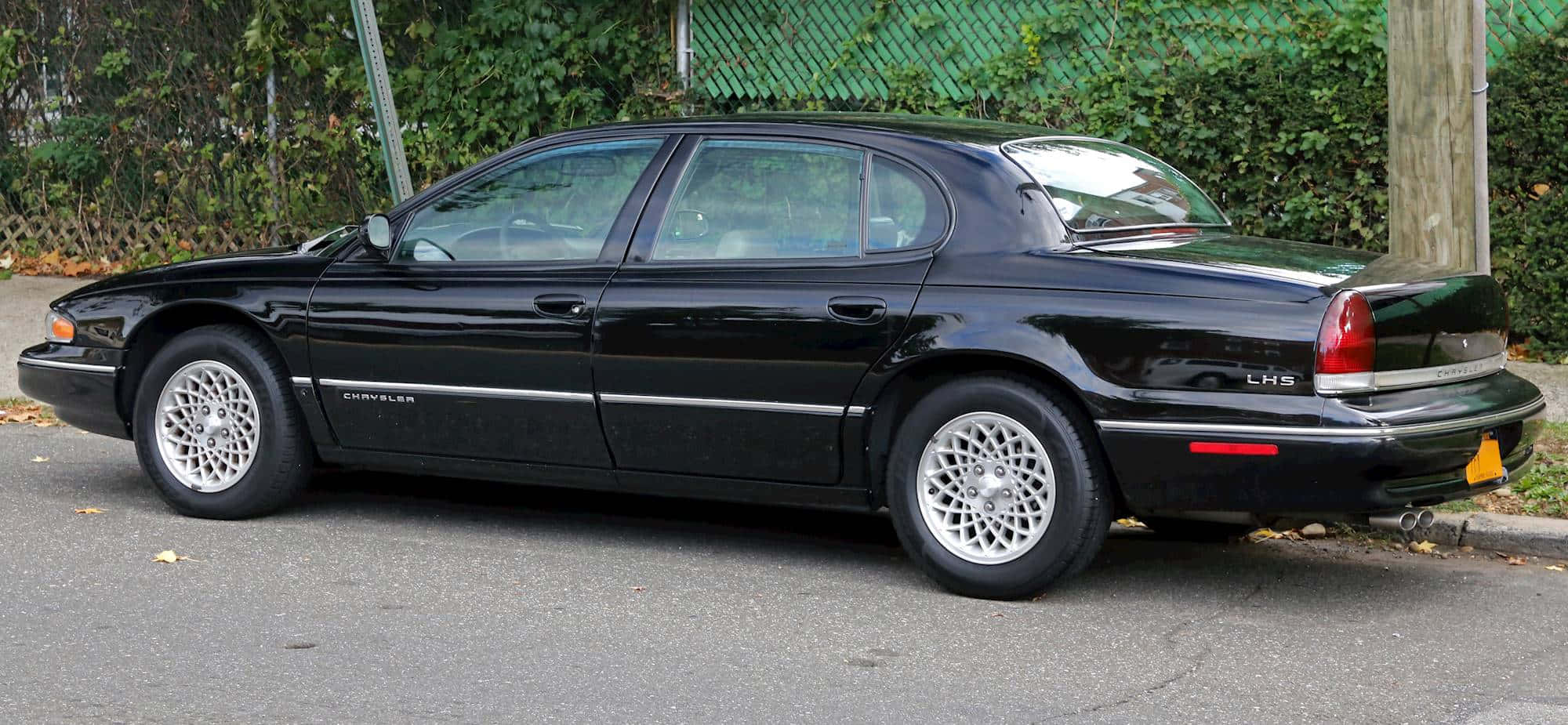 Chrysler Lhs 桌布 壁紙 — 一輛時尚的黑色克萊斯勒LHS轎車停在街上，旁邊是綠色鐵絲網圍欄和茂密的灌木叢。