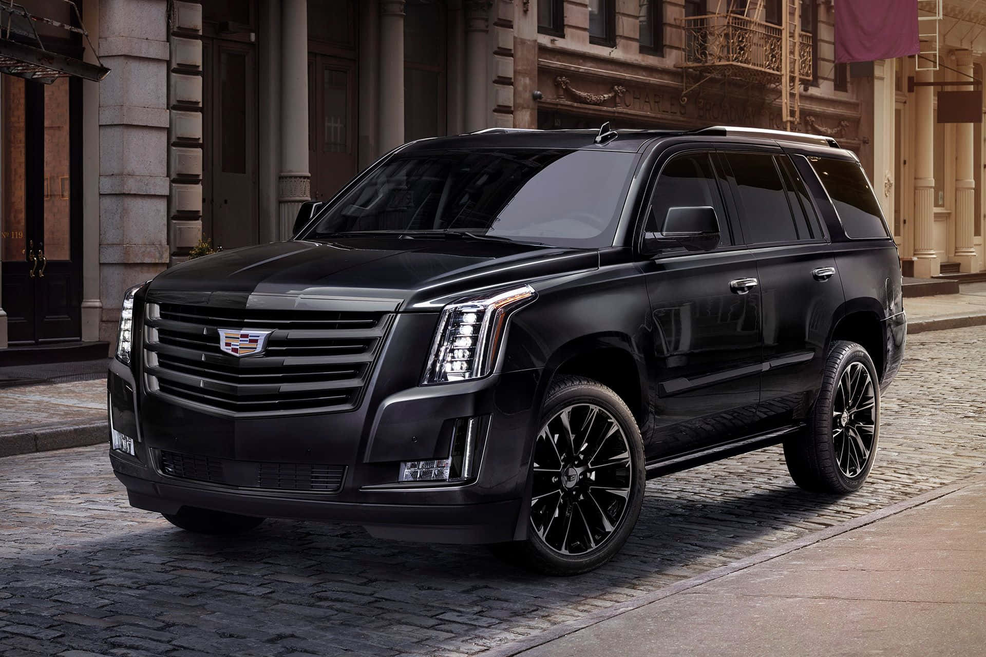 Family Car 桌布 壁紙 — 一輛時尚的黑色凱迪拉克 Escalade SUV 停放在歷史石砌建築前的鵝卵石街道上。
