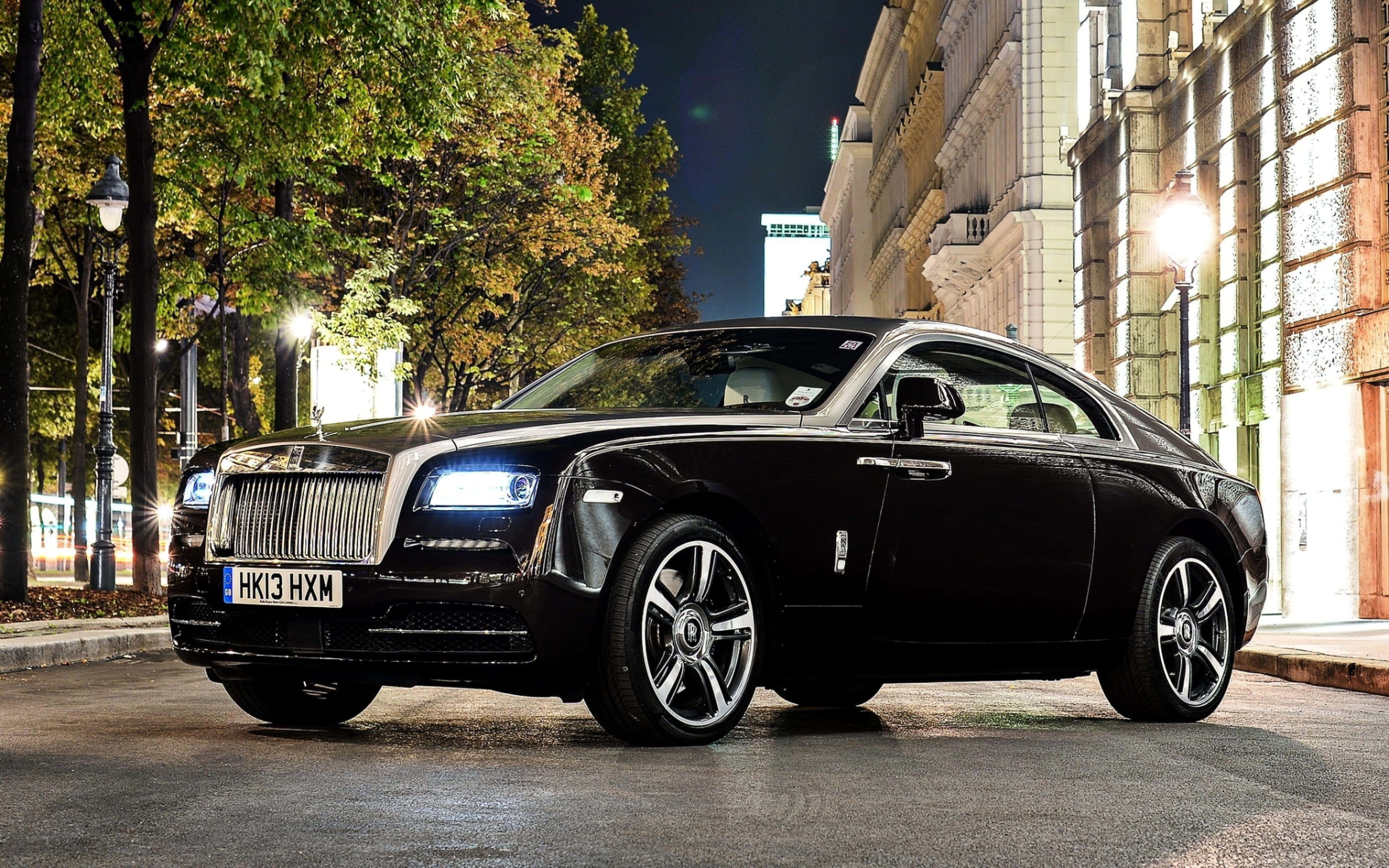 Rolls Royce 桌布 壁紙 — 一輛時尚的黑色勞斯萊斯魅影在城市燈光下閃耀，停在濕漉漉的秋日街道上，兩旁種滿樹木和歷史建築。