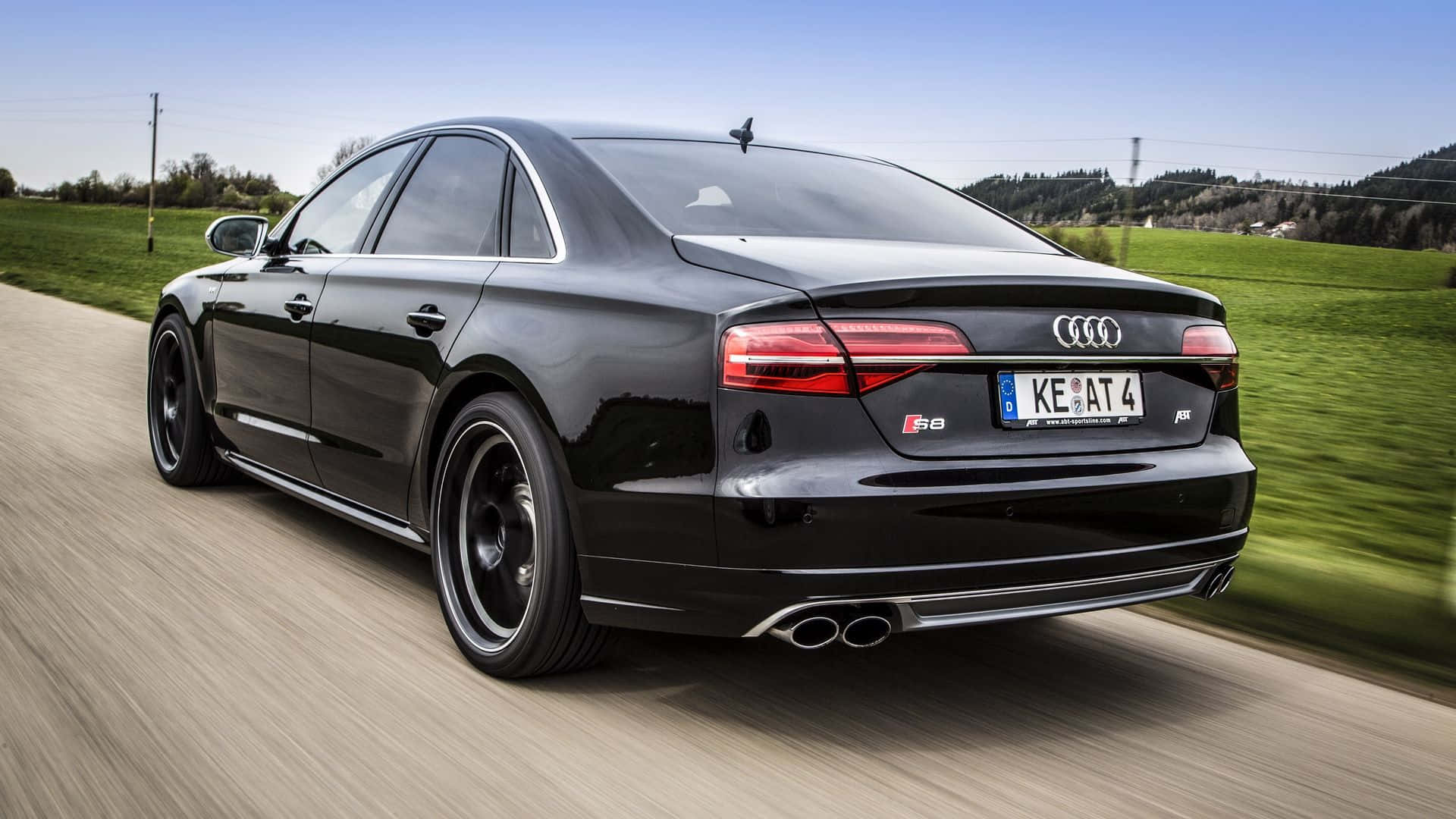 Audi S8 桌布 壁紙 — 一輛時尚的黑色奧迪S8轎車在鄉村道路上飛馳，背景是模糊的綠色田野和山丘，頭頂是清澈的藍天。