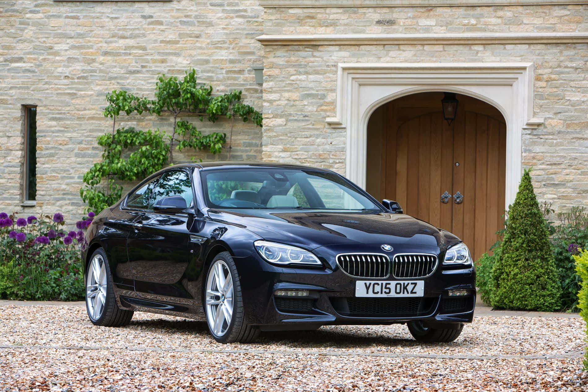 Bmw 6 Series 桌布 壁紙 — 一輛時尚的黑色寶馬6系轎跑車停在碎石路上，前方是一座帶有拱形木門和攀緣植物的石砌莊園。