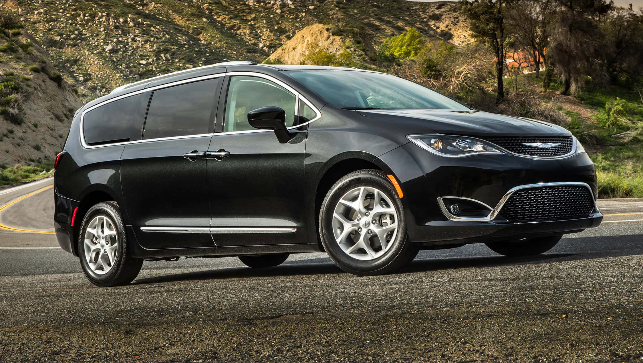 Chrysler Pacifica 桌布 壁紙 — 一輛時尚的黑色小型客車在明亮的日光下行駛在一條蜿蜒的山路上，周圍是岩石地形和稀疏的綠植。