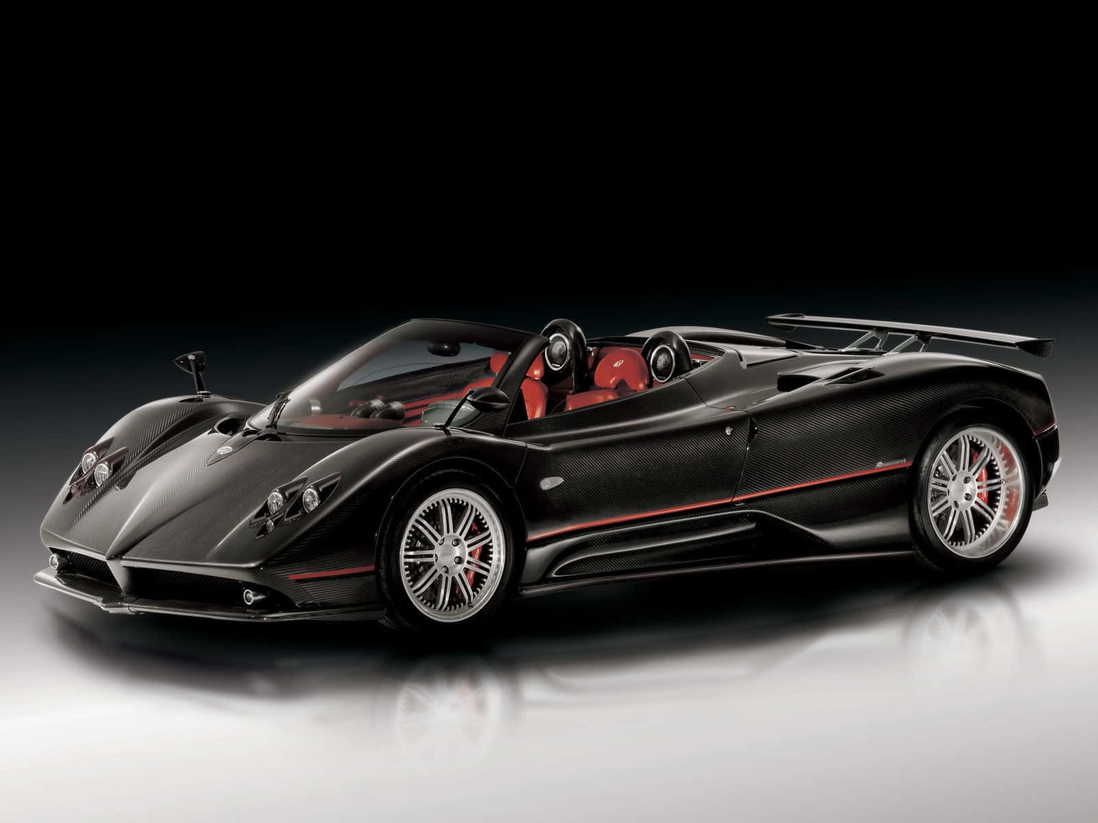 Pagani Zonda C12 Roadster 桌布 壁紙 — 一輛時尚的黑色帕加尼Zonda敞篷跑車，配備紅色內飾和碳纖維細節，在深色背景下的反光工作室地板上滑行。