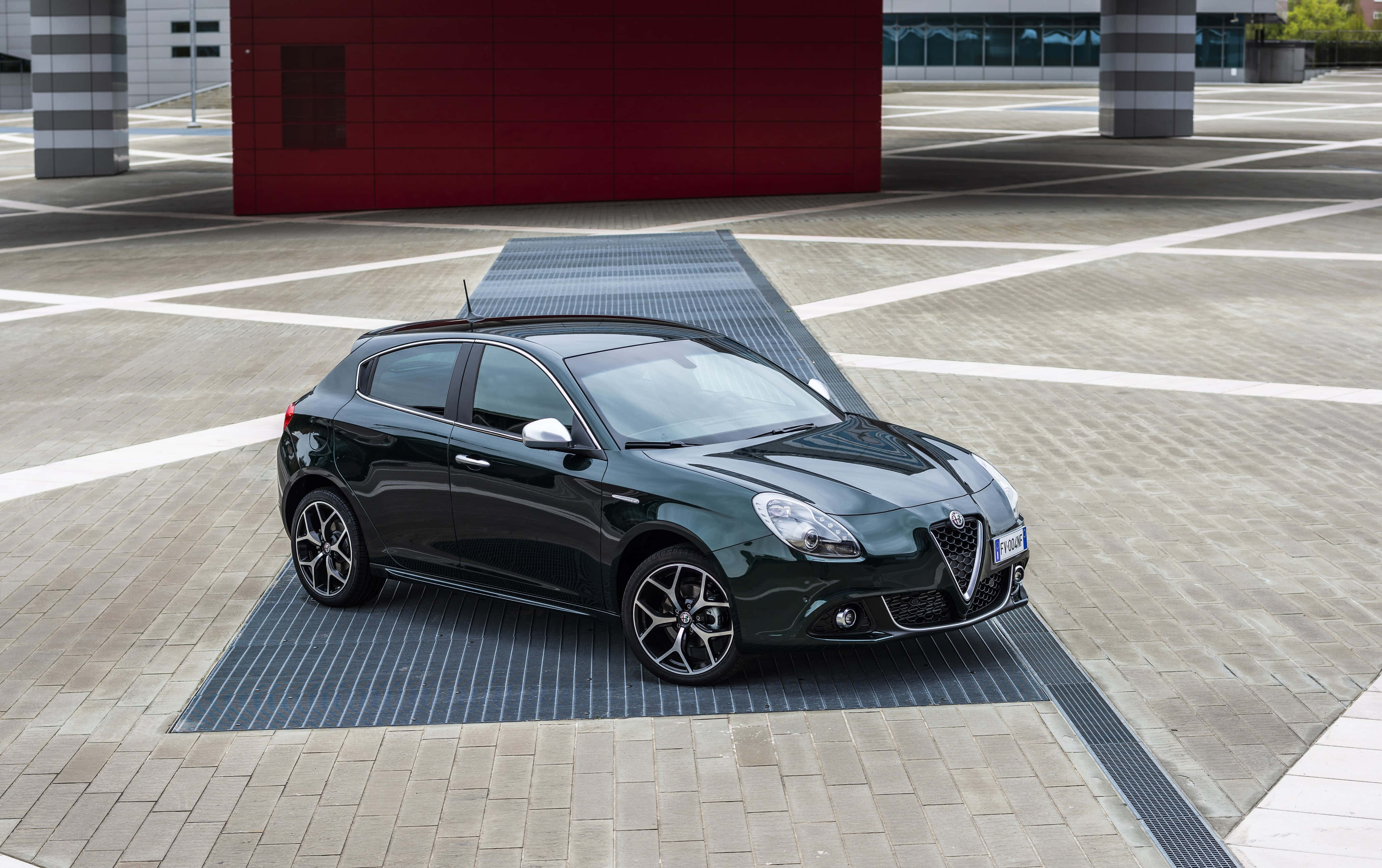 Alfa Romeo Giulietta 桌布 壁紙 — 一輛時尚的黑色愛快羅密歐 Giulietta 停泊在現代鋪砌廣場上，背景是鮮明的紅色建築。