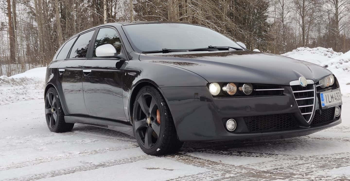 Alfa Romeo 159 桌布 壁紙 — 一輛時尚的黑色愛快羅密歐旅行車停泊在積雪的森林道路上，背景是光禿禿的樹木。