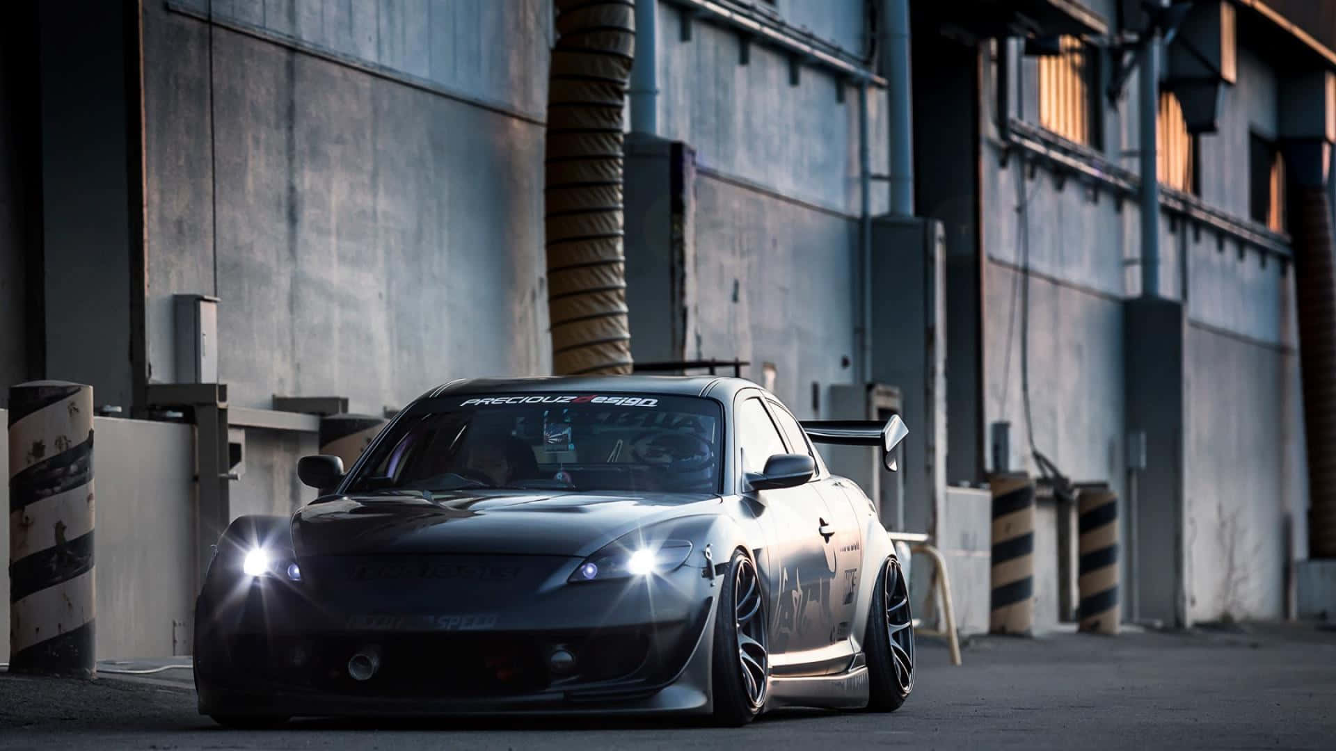 Mazda Rx-8 桌布 壁紙 — 一輛時尚的黑色改裝跑車，帶有發光的車頭燈，在黃昏時停放在工業小巷中。