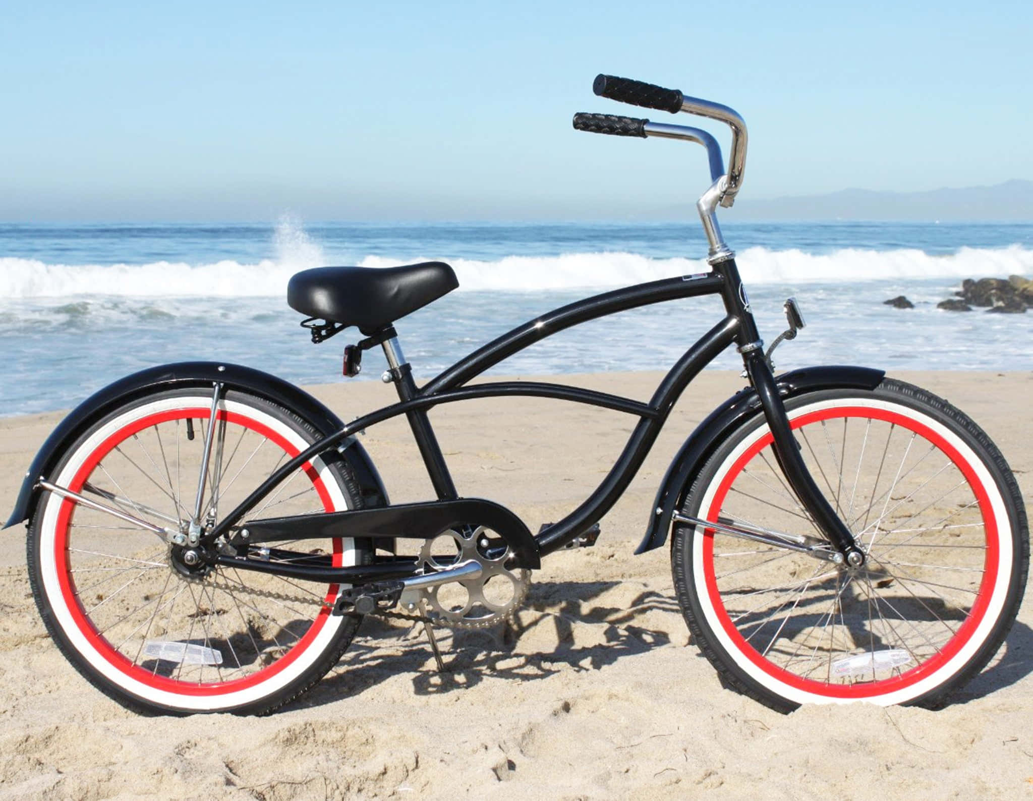 Beach Cruiser 桌布 壁紙 — 一輛時尚的黑色沙灘巡航自行車，配有紅色和白色輪胎，停放在陽光普照的沙灘上，旁邊是拍打的海浪。