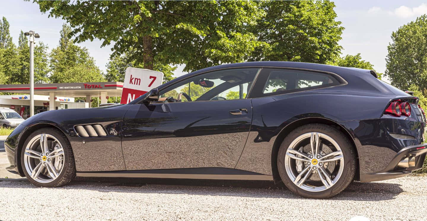 Ferrari Gtc4lusso 桌布 壁紙 — 一輛時尚的黑色法拉利 GTC4Lusso 停在加油站旁的碎石地上，周圍有茂密的綠樹，在明亮的日光下。