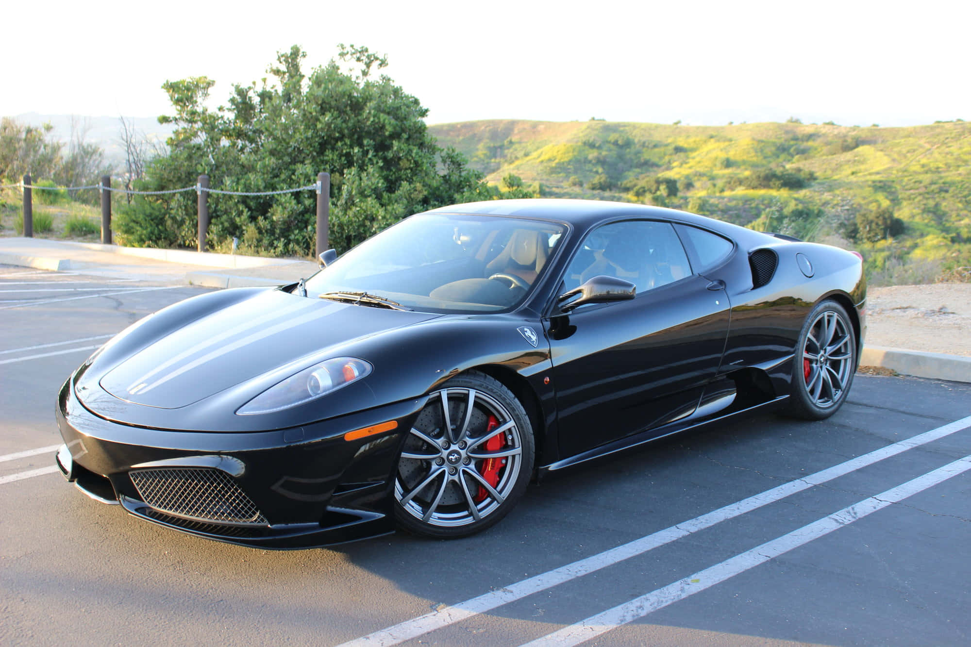 Ferrari F430 桌布 壁紙 — 一輛時尚的黑色法拉利跑車，配備紅色制動卡鉗，停泊在陽光普照的停車場，背景是起伏的綠色山丘。