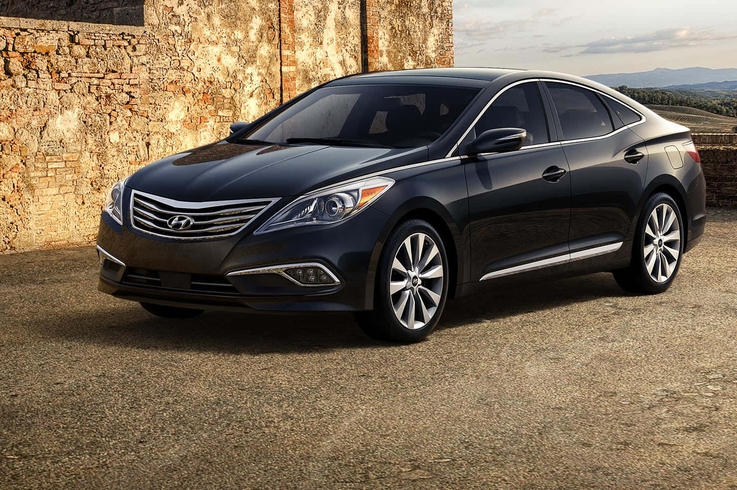 Hyundai Azera 桌布 壁紙 — 一輛時尚的黑色現代轎車停在碎石路上，旁邊是風化的石牆，遠處有山丘，天空柔和。