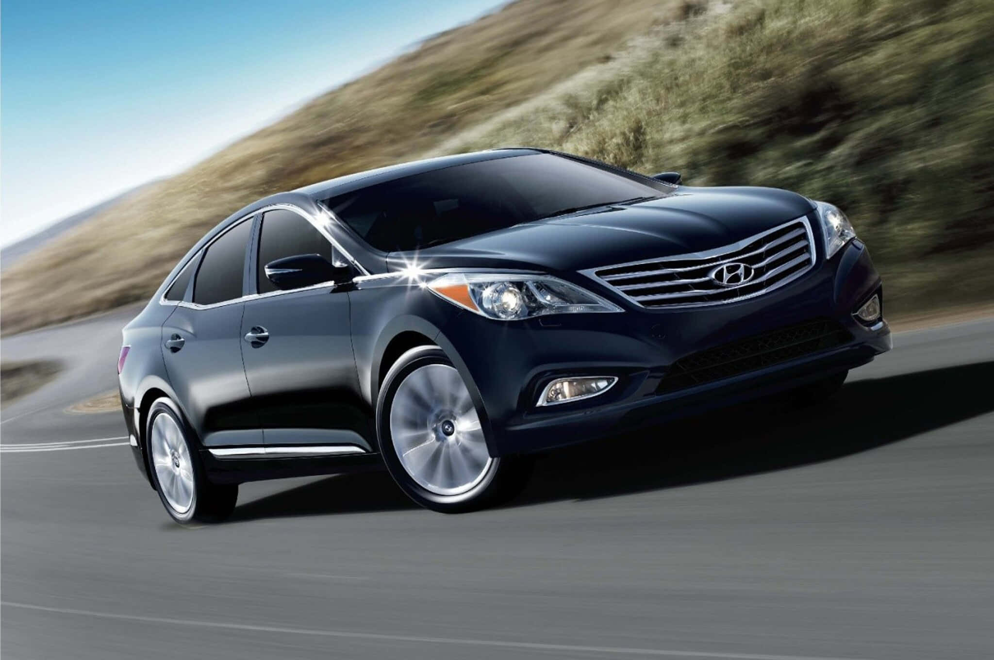Hyundai Azera 桌布 壁紙 — 一輛時尚的黑色現代轎車在蜿蜒的道路上行駛，背景是模糊的山丘，天空清澈湛藍。