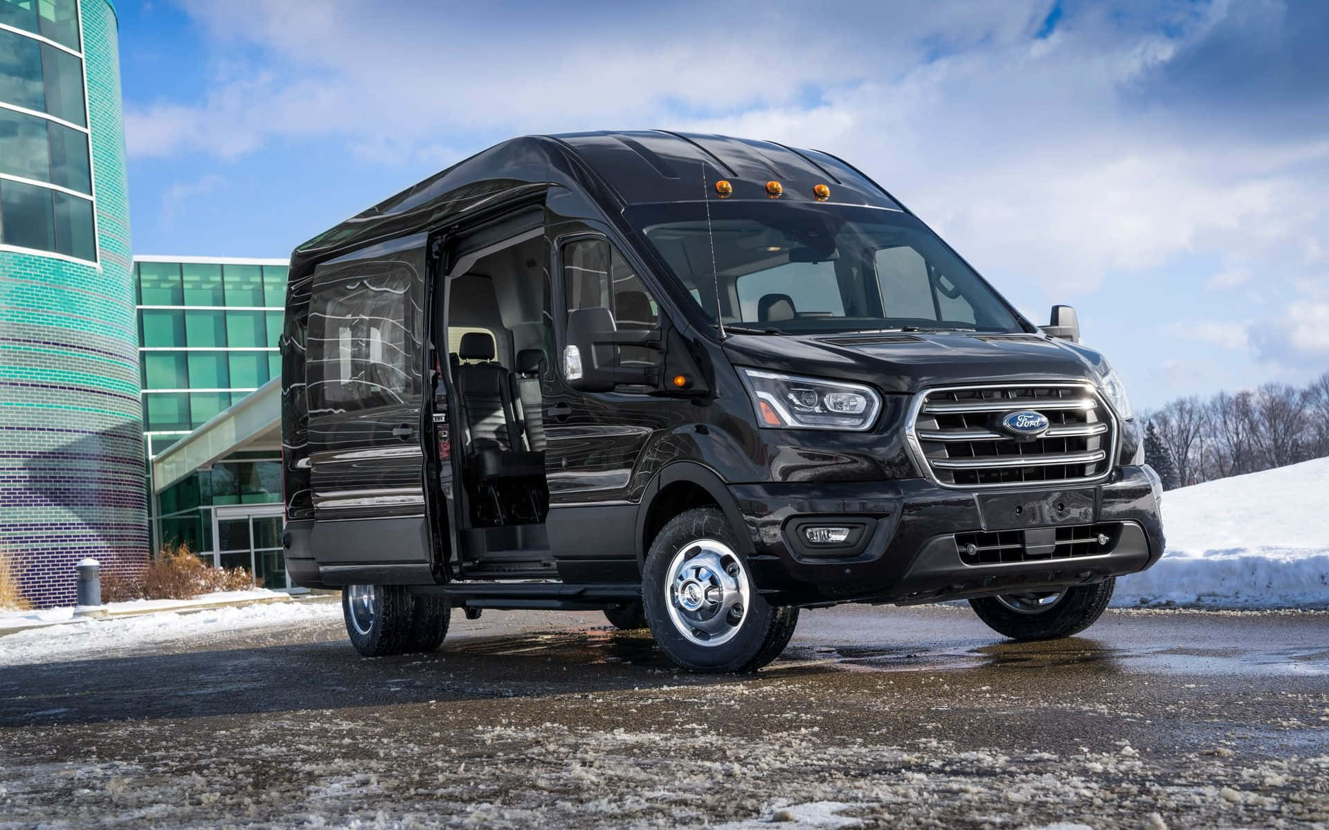 Ford Transit 桌布 壁紙 — 一輛時尚的黑色福特露營車停泊在雪地上，旁邊是一座現代玻璃建築，上方是明亮的藍天。