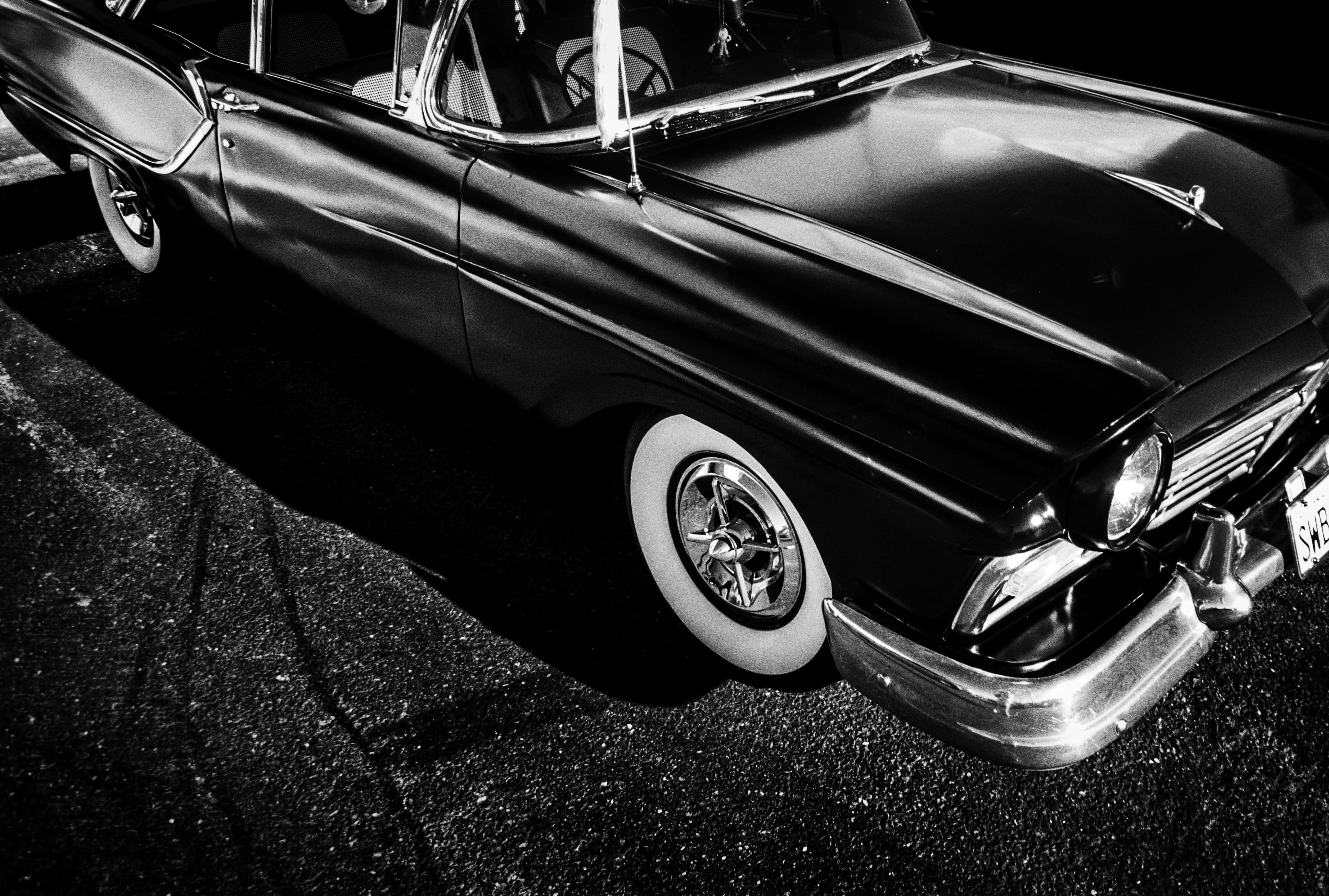 Black And White Vintage Car 桌布 壁紙 — 一輛時尚的黑色經典汽車在戲劇性的燈光下閃閃發光，在瀝青上投下銳利的陰影。