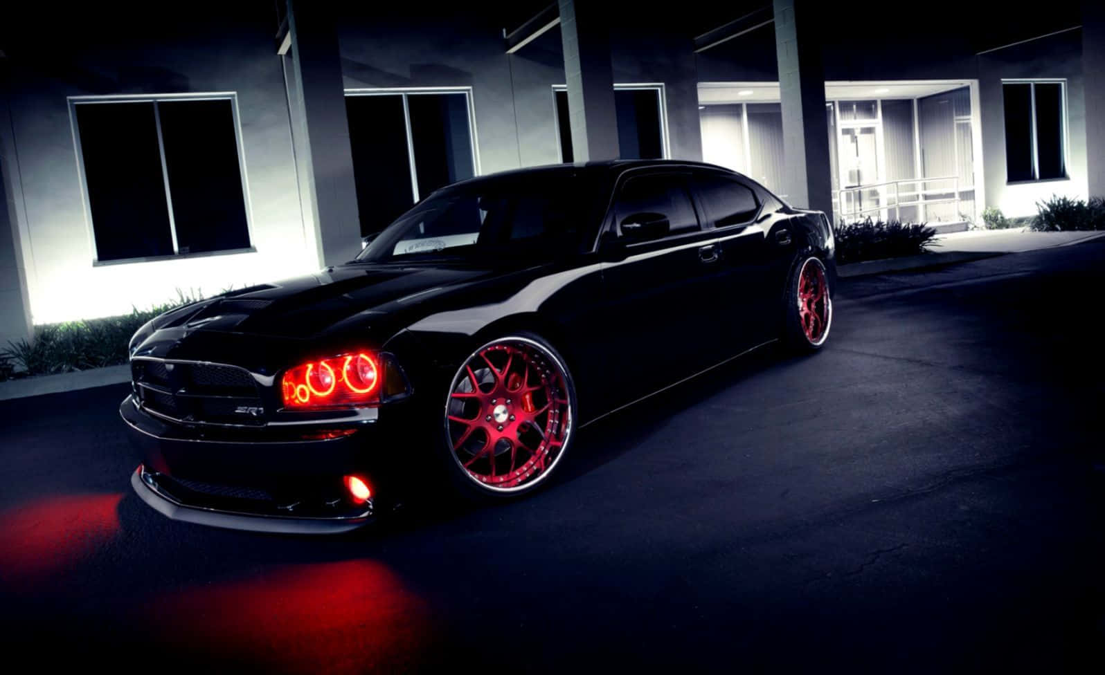 Dodge Charger 桌布 壁紙 — 一輛時尚的黑色肌肉車，帶有發光的紅色輪圈和頭燈，夜間停在一棟現代建築前。