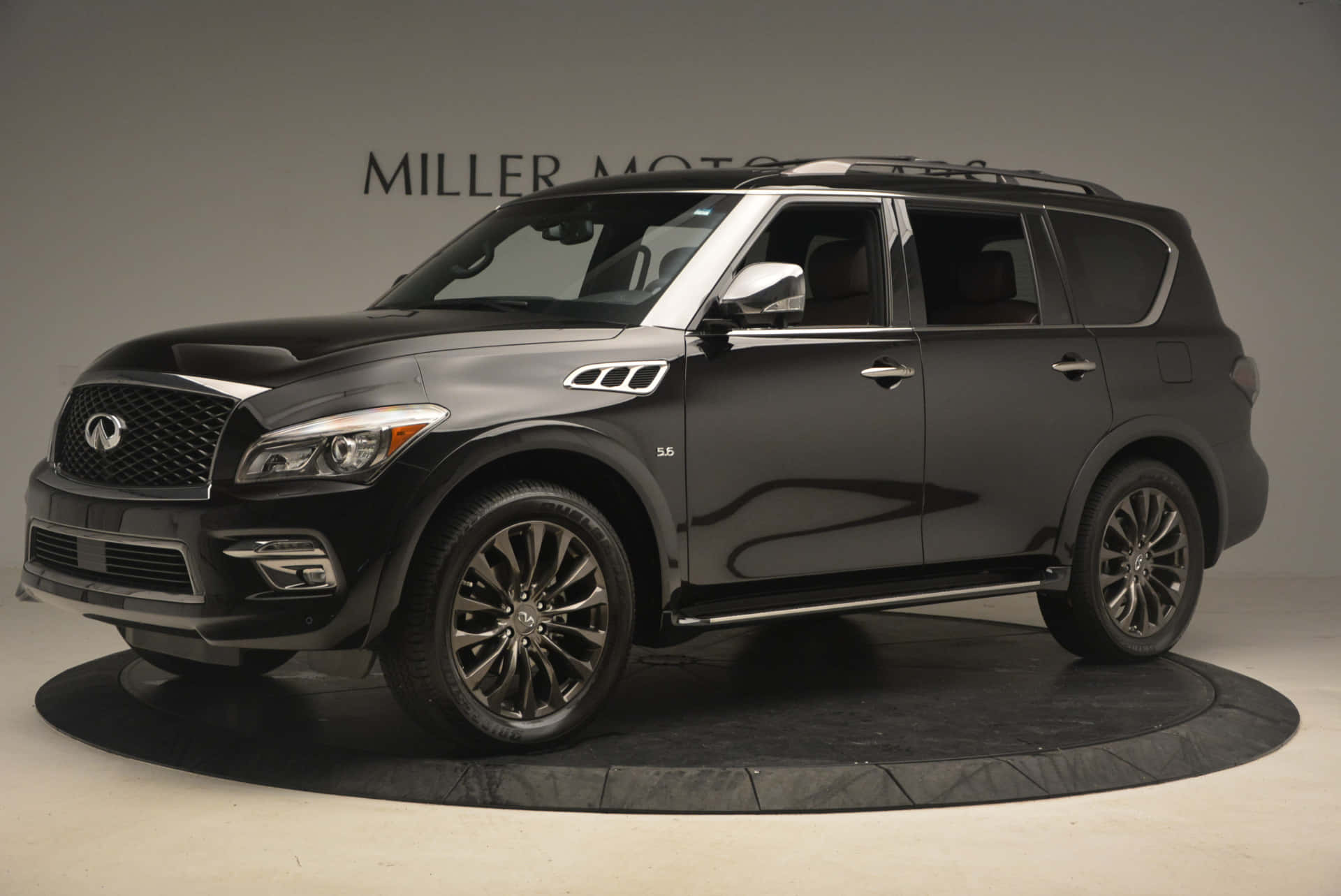 Infiniti Qx80 桌布 壁紙 — 一輛時尚的黑色英菲尼迪 QX80 SUV 在極簡主義工作室的圓形平台上展示，牆上帶有 'MILLER MOTORS' 品牌標識。