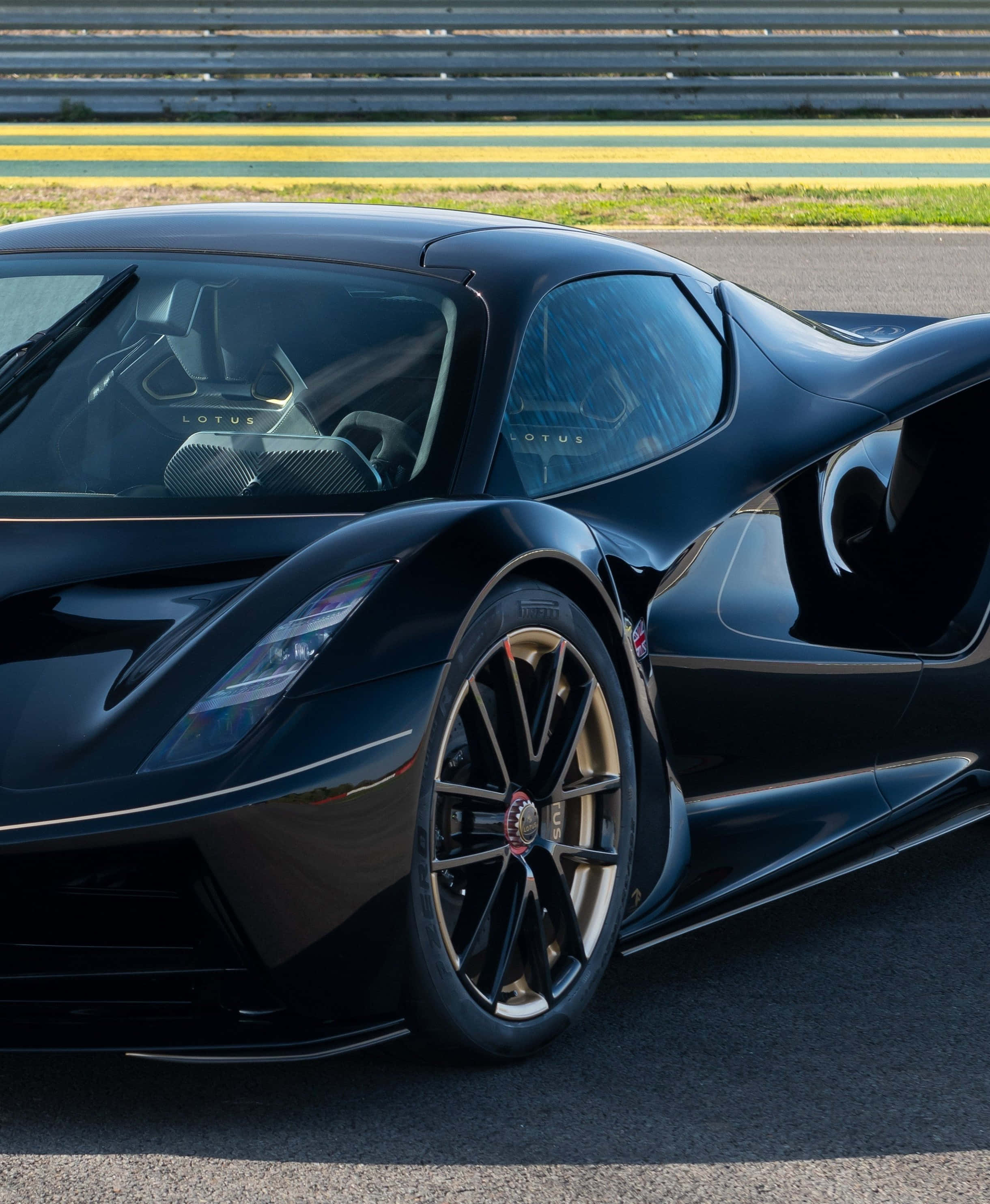 Sports Car 桌布 壁紙 — 一輛時尚的黑色蓮花超級跑車在明亮的陽光下，在帶有黃色路緣和金屬護欄的賽道上閃閃發光。