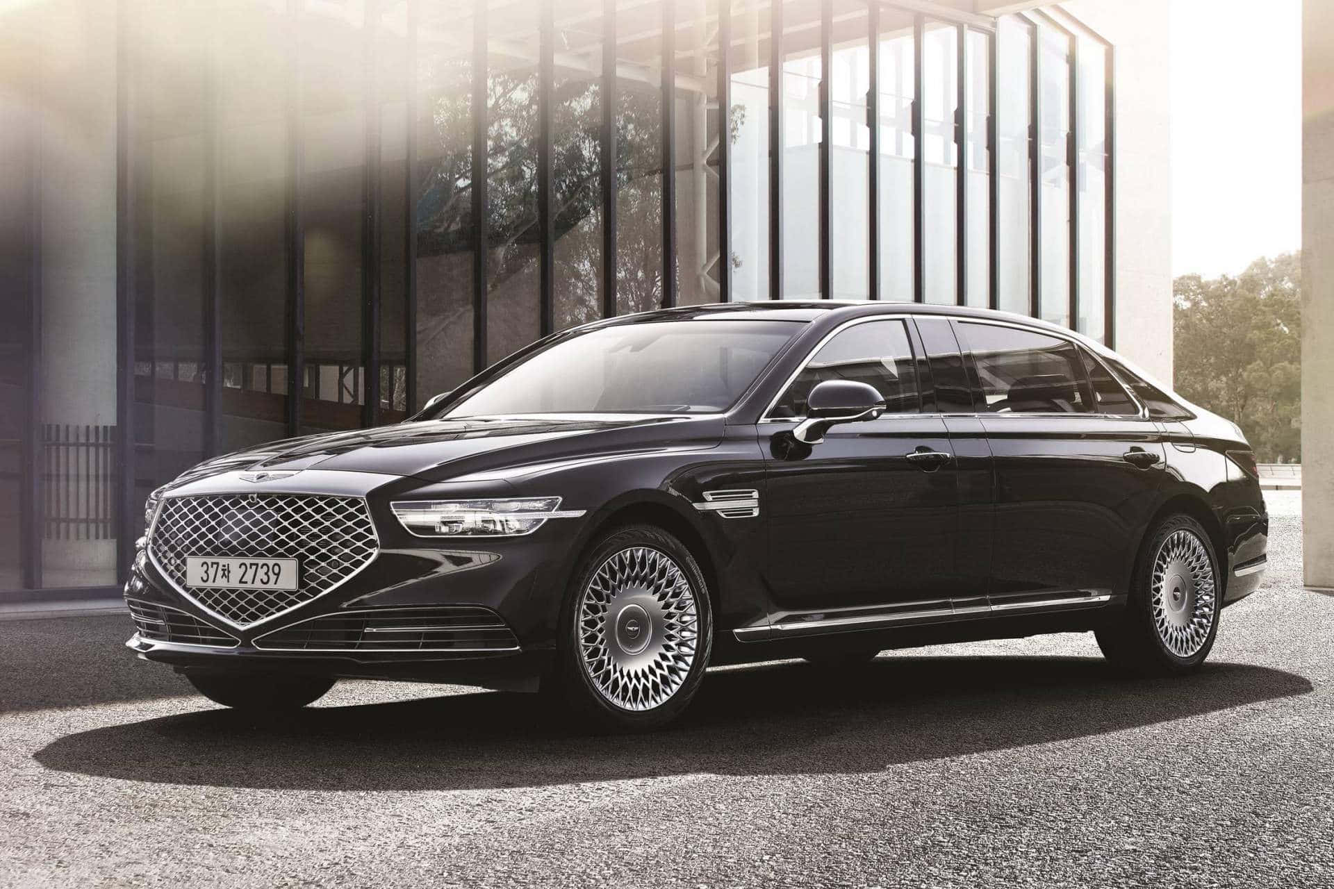 Genesis G90 桌布 壁紙 — 一輛時尚的黑色豪華轎車，帶有複雜的輪圈，停在現代玻璃建築前，陽光透過建築照射進來。