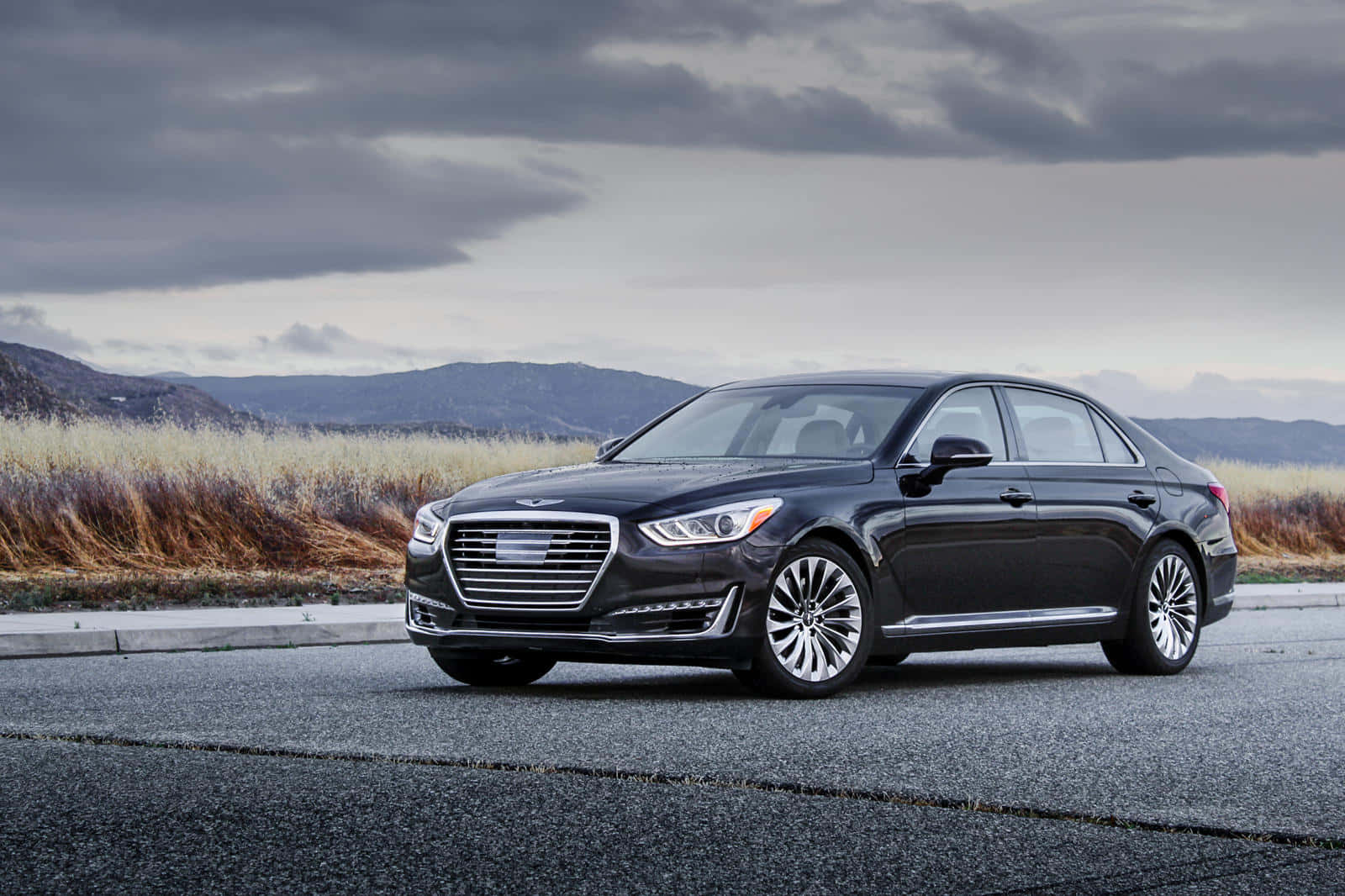 Genesis G90 桌布 壁紙 — 一輛時尚的黑色豪華轎車停在瀝青路上，周圍有乾草和遠山，天空多雲。