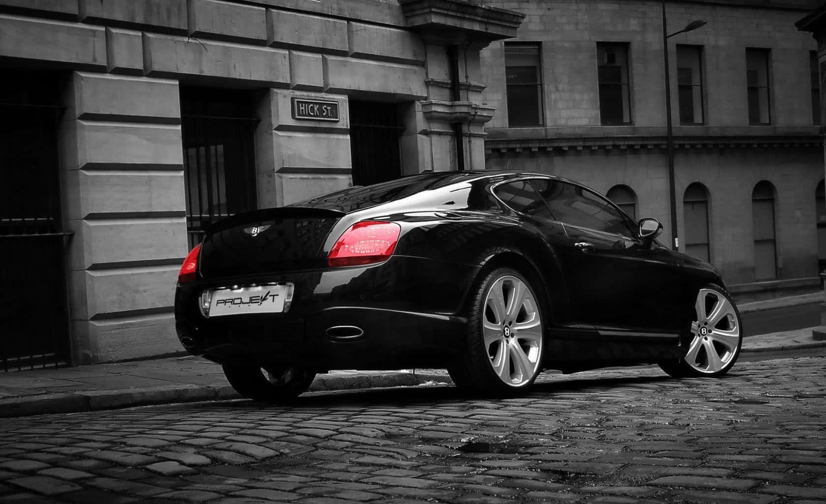 Bentley Continental Gt 桌布 壁紙 — 一輛時尚的黑色賓利 Continental GT 停泊在鵝卵石街道上，在單色城市背景下，尾燈的紅色透過選擇性色彩效果突出顯示。