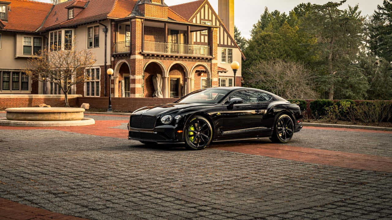 Bentley Continental Gt 桌布 壁紙 — 一輛時尚的黑色賓利 Continental GT，帶有黃色制動卡鉗，停在鵝卵石車道上，前方是一座宏偉的都鐸風格豪宅。