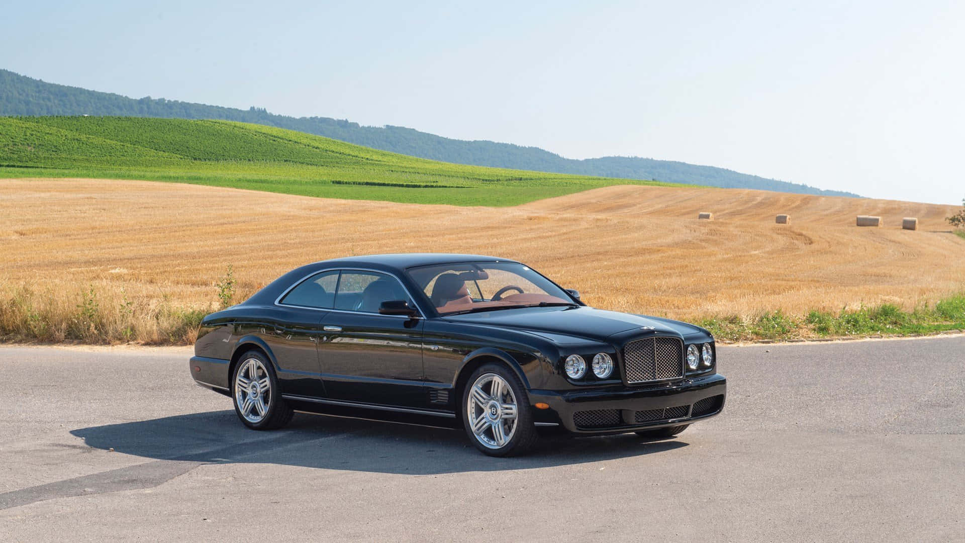 Bentley Brooklands 桌布 壁紙 — 一輛時尚的黑色賓利 Continental GT 停泊在鄉村道路上，周圍是金黃色的田野和翠綠的山丘，天空清澈湛藍。