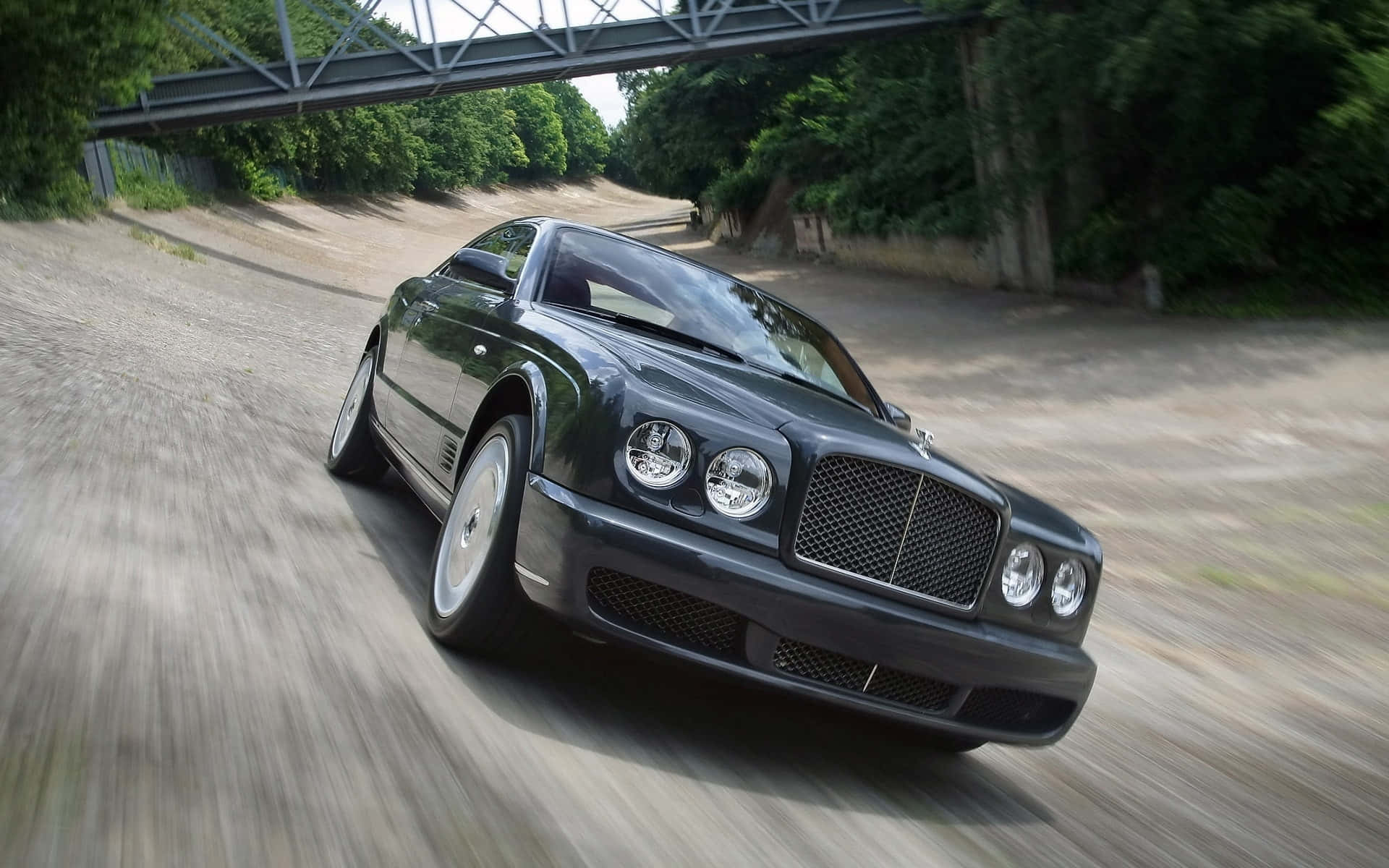 Bentley Brooklands 桌布 壁紙 — 一輛時尚的黑色賓利在鋼橋下的碎石路上疾馳，周圍是茂密的綠樹。