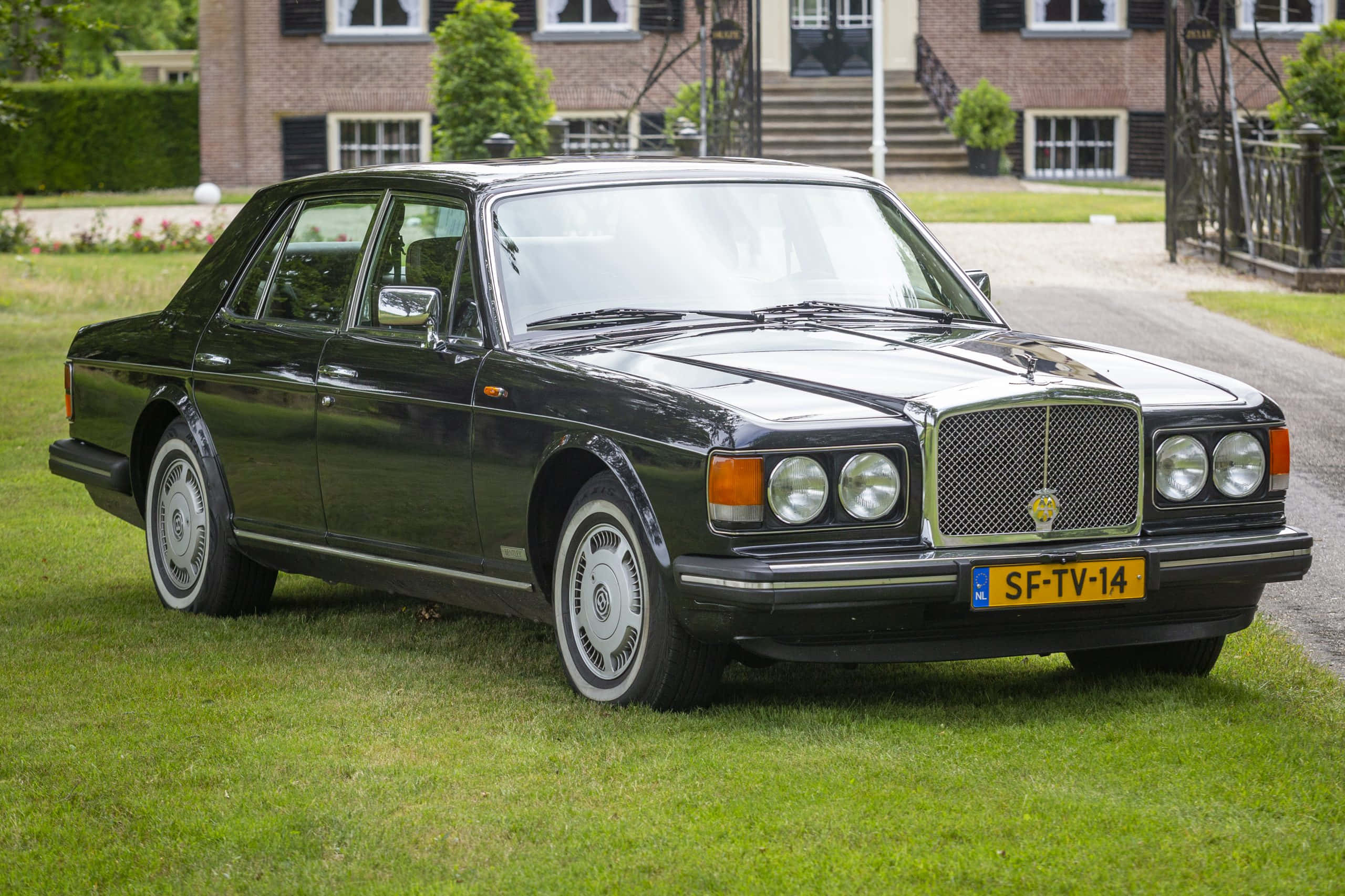 Bentley Eight 桌布 壁紙 — 一輛時尚的黑色賓利轎車停泊在生氣勃勃的綠色草地上，前面是一座莊嚴的磚砌豪宅，配有修剪整齊的樹籬。