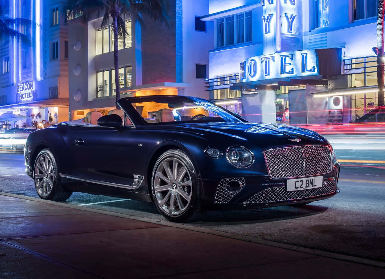 Bentley Continental Gt Convertible 桌布 壁紙 — 一輛時尚的黑色賓利開篷車在夜晚停泊在霓虹燈裝飾的Art Deco酒店前，周圍有棕櫚樹和模糊的城市燈光。