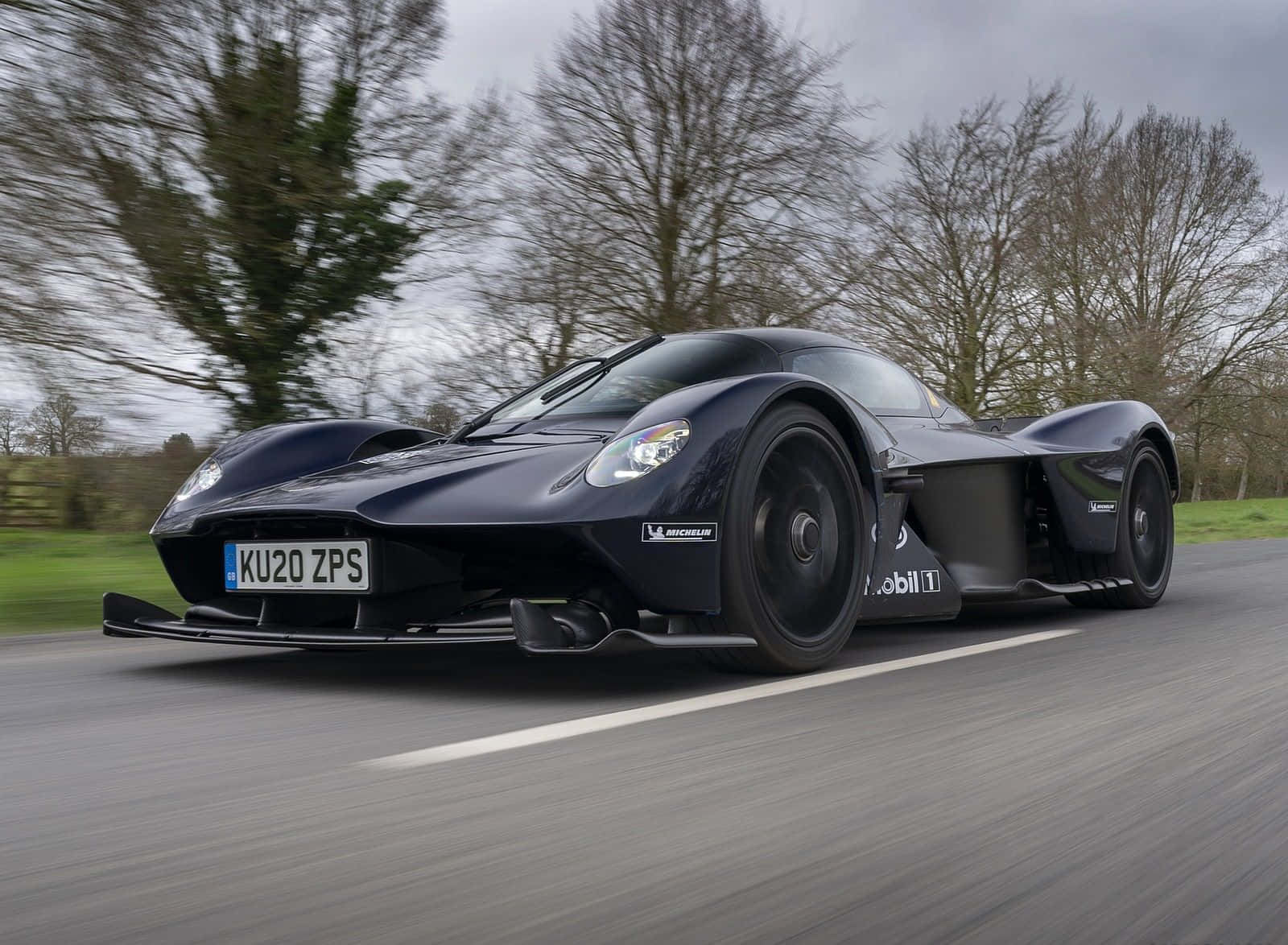 Aston Martin Valkyrie 桌布 壁紙 — 一輛時尚的黑色超跑在鄉村道路上疾馳，背景是模糊的樹木和陰天。