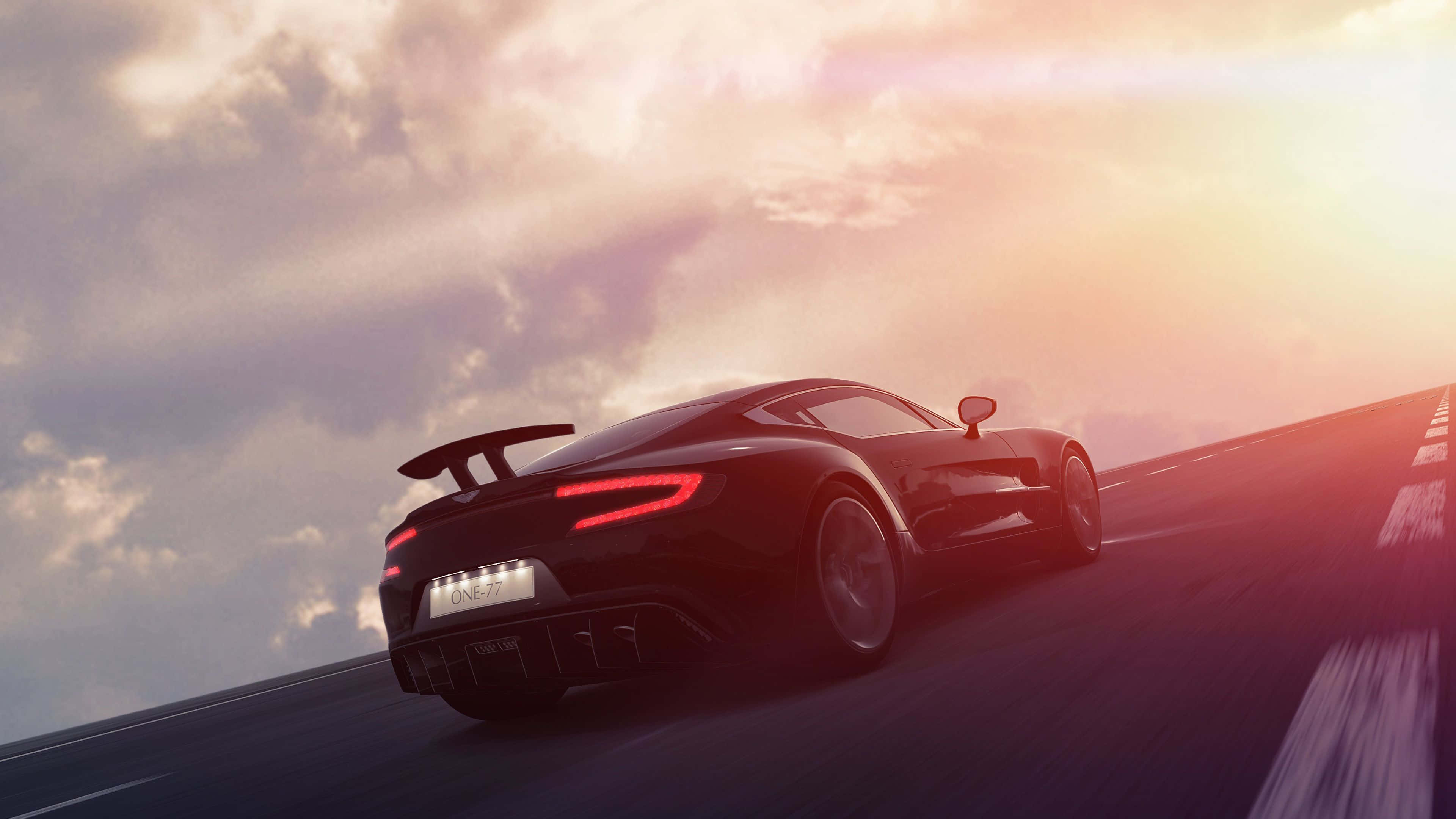 Aston Martin One-77 桌布 壁紙 — 一輛時尚的黑色跑車，帶有發光的紅色尾燈，在戲劇性的天空下沿著蜿蜒的道路在日落時分疾馳。