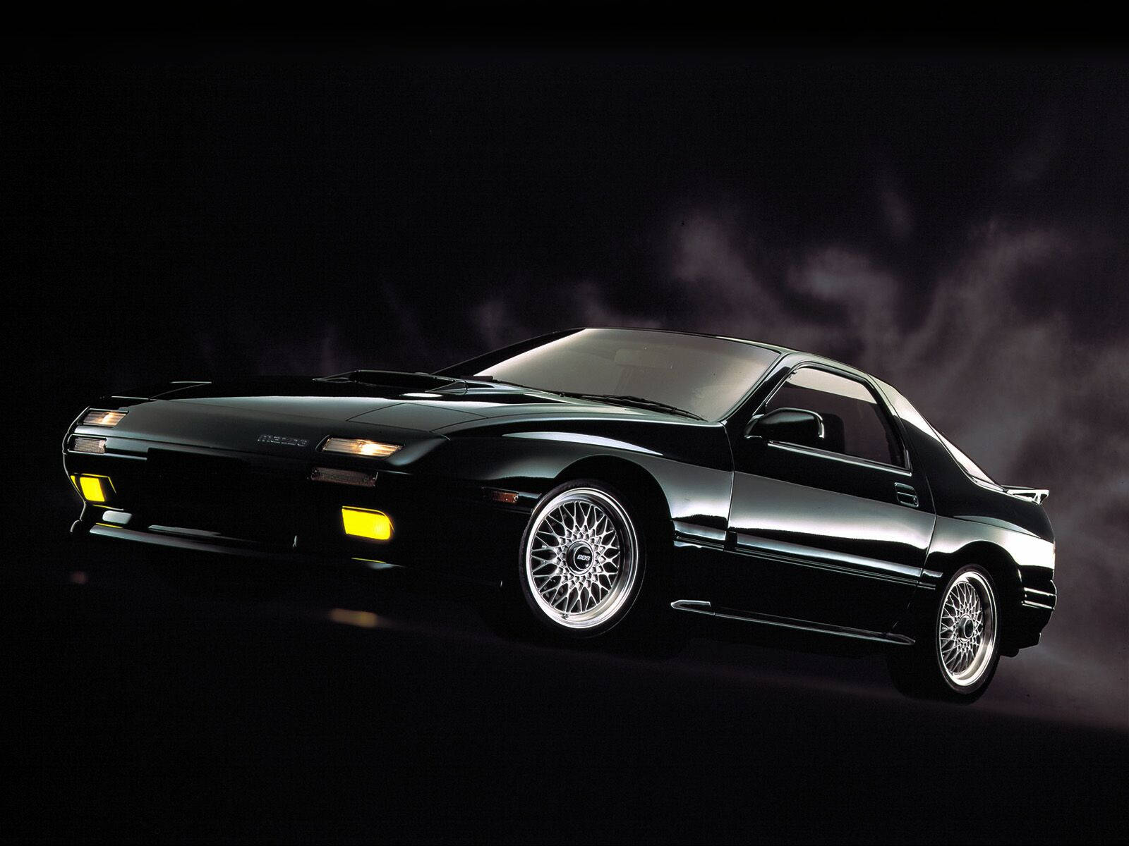 Mazda 桌布 壁紙 — 一輛時尚的黑色跑車，帶有發光的黃色頭燈和銀色輪圈，在煙霧瀰漫的黑暗背景下戲劇性地照亮。