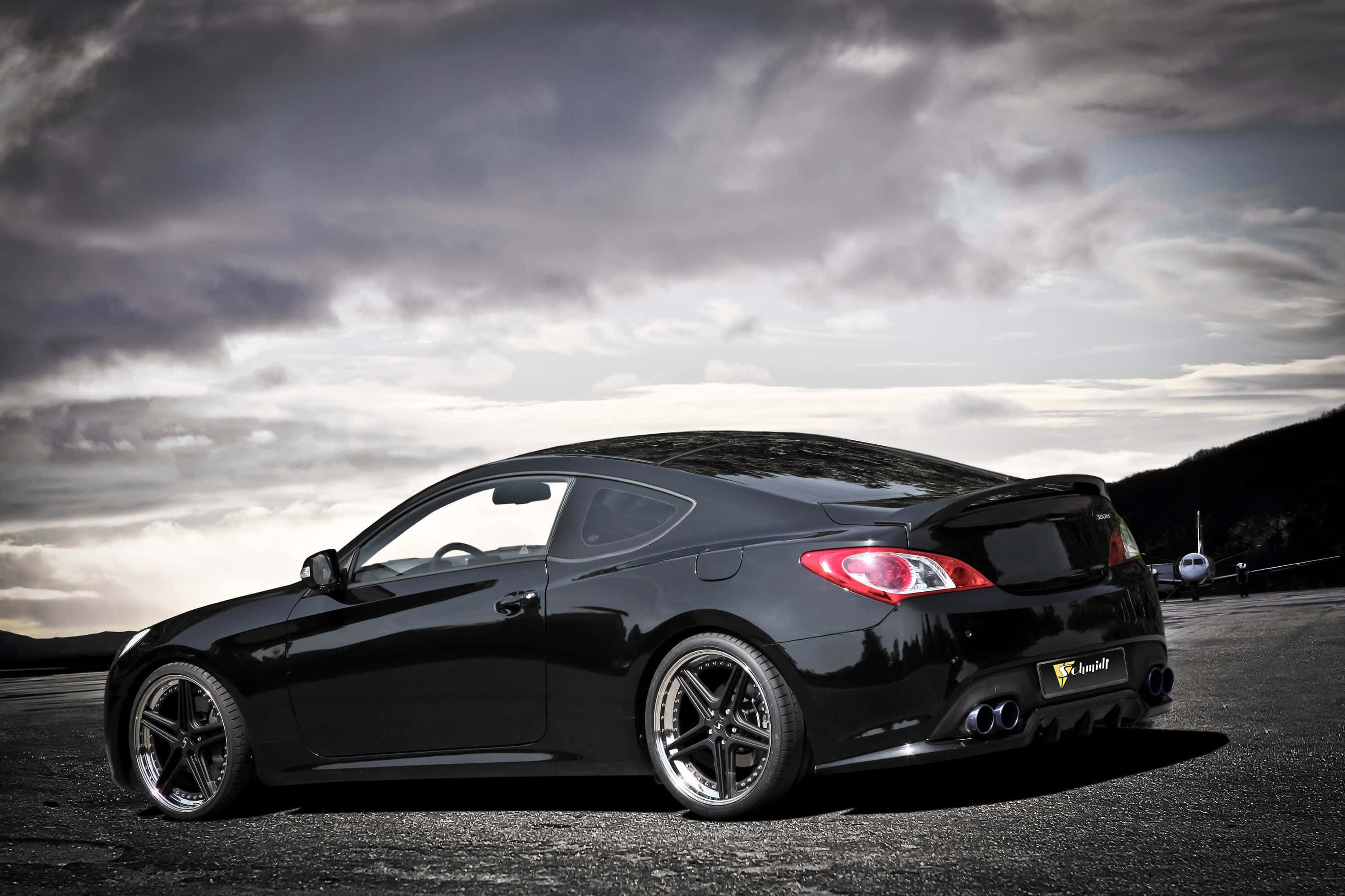 Hyundai Genesis 桌布 壁紙 — 一輛時尚的黑色跑車，配備定制輪轂和雙排氣管，停在戲劇性多雲天空下的跑道上。