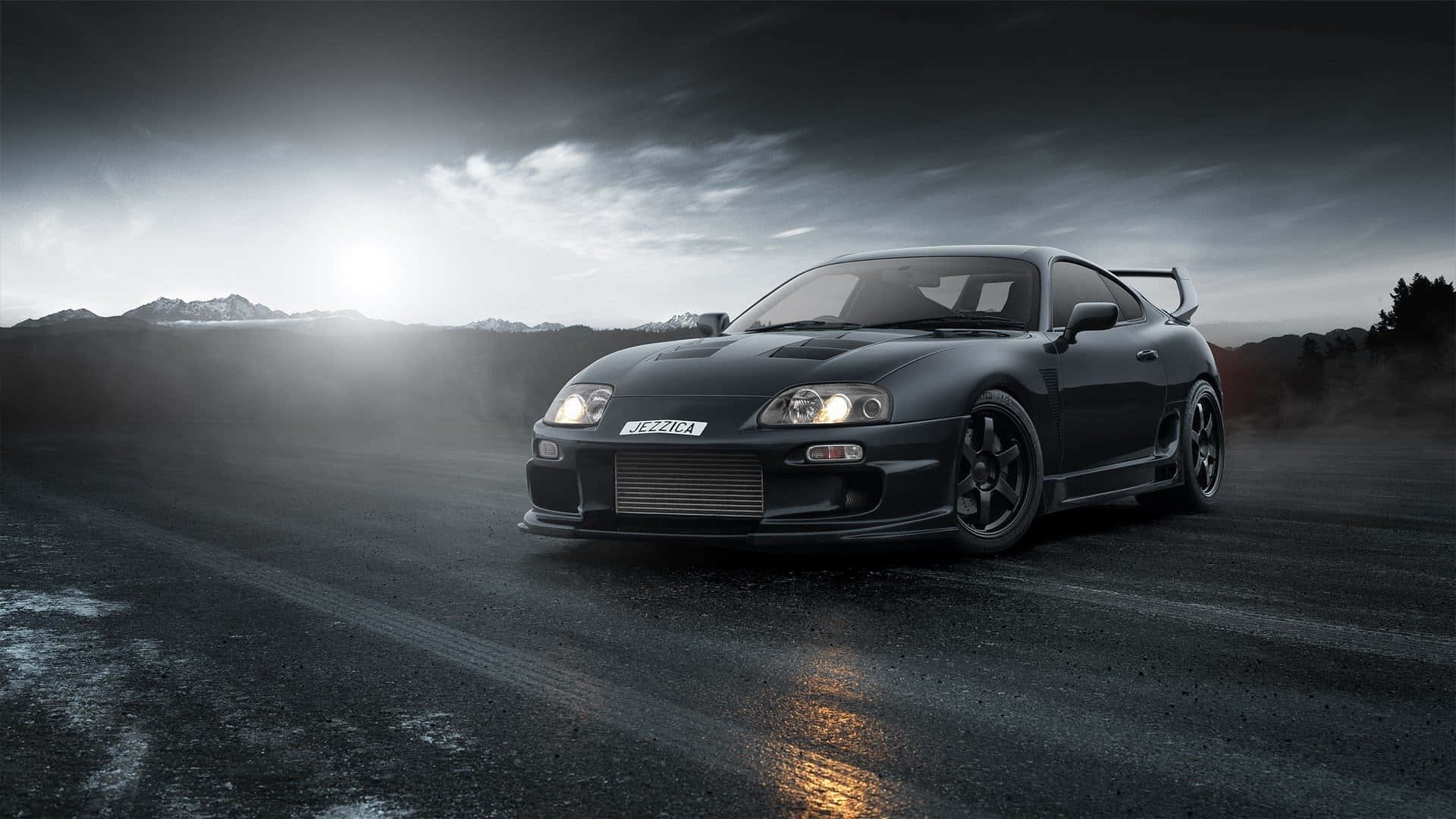 Supra 桌布 壁紙 — 一輛時尚的黑色跑車，配備發光頭燈，在潮濕的道路上奔馳，背景是戲劇性的多雲天空和遠山。