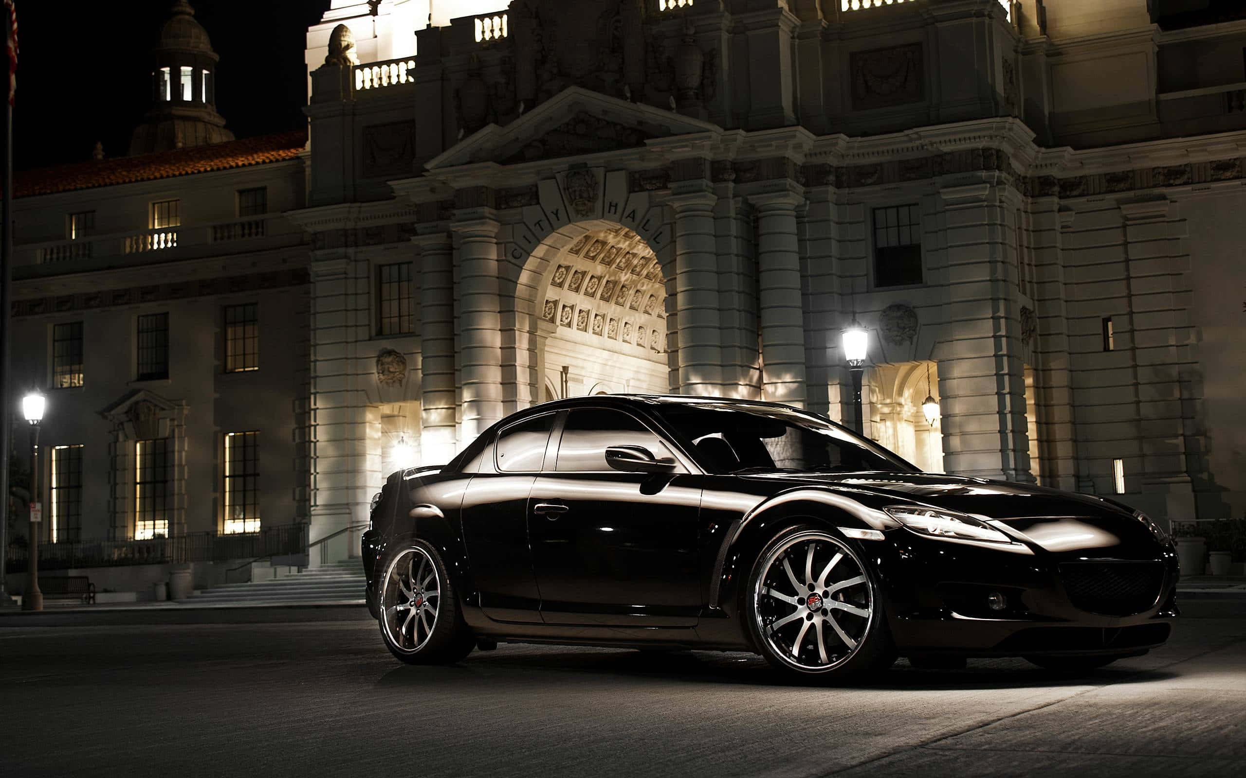 Mazda Rx-8 桌布 壁紙 — 一輛時尚的黑色跑車在夜晚的街燈下閃閃發光，停在一座宏偉的燈火通明的新古典主義建築前。